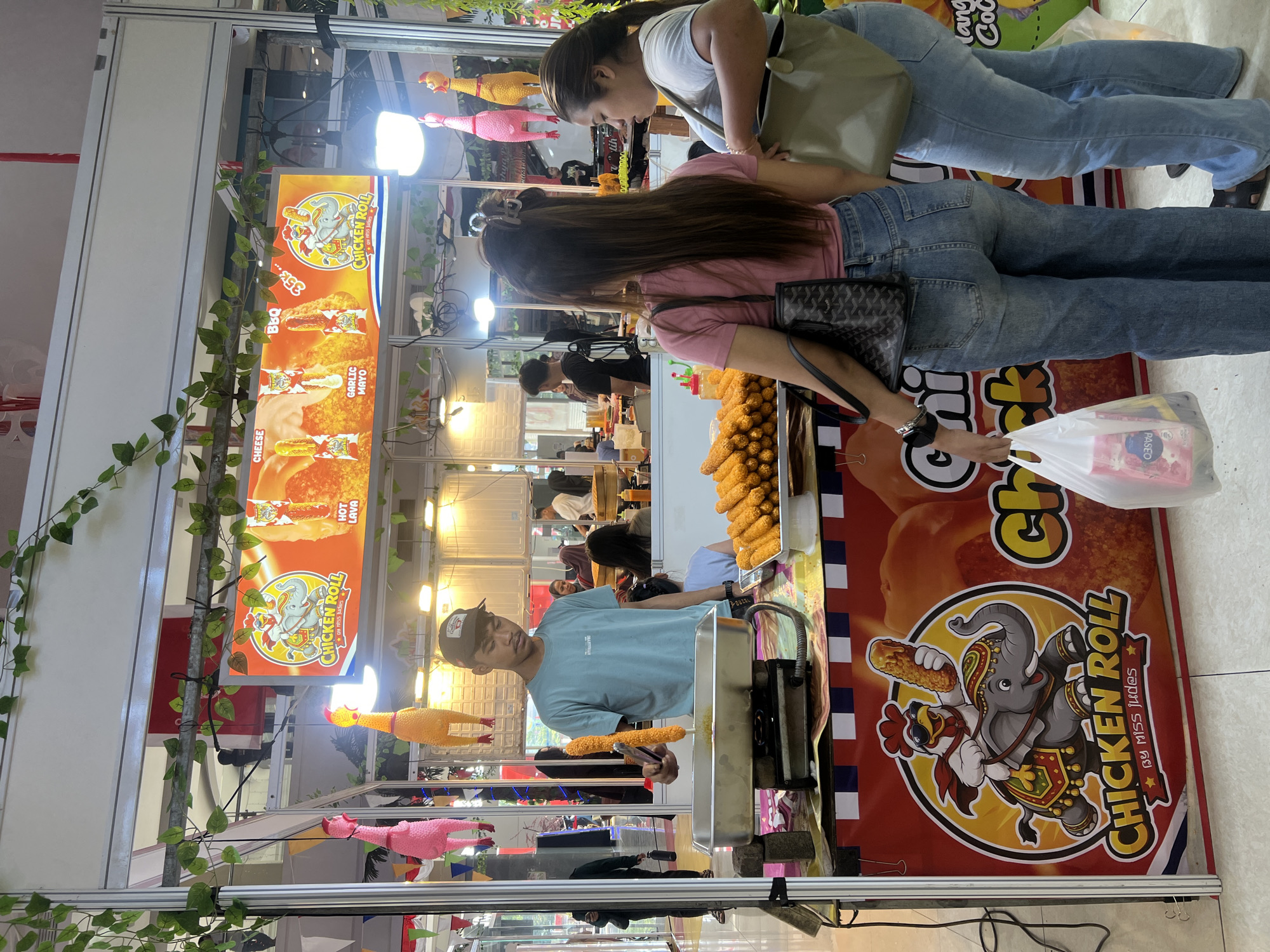 The image depicts an outdoor food stall in Banyurojo, Indonesia, specializing in "Chicken Roll." A young man, wearing a light blue t-shirt and a dark baseball cap, is actively preparing food. He holds a fried chicken product with tongs over a metal tray, which sits on a portable stove. In the foreground, a large display features a colorful banner with an animated chicken riding an elephant, promoting "Chicken Roll by Miss Judes." Piles of freshly fried chicken rolls are arranged on a white platter to the right. Two women are visible, facing away from the camera and observing the stall. The woman on the left wears a pink t-shirt and denim jeans, carrying a black handbag and a white plastic bag containing packaged goods. The woman on the right is in light blue jeans and a light-colored top, carrying a large green tote bag. Overhead, decorative green vines and pink and yellow hanging toys shaped like chickens add to the festive atmosphere of the stall. The background reveals more of the marketplace, including other stalls and a few patrons.