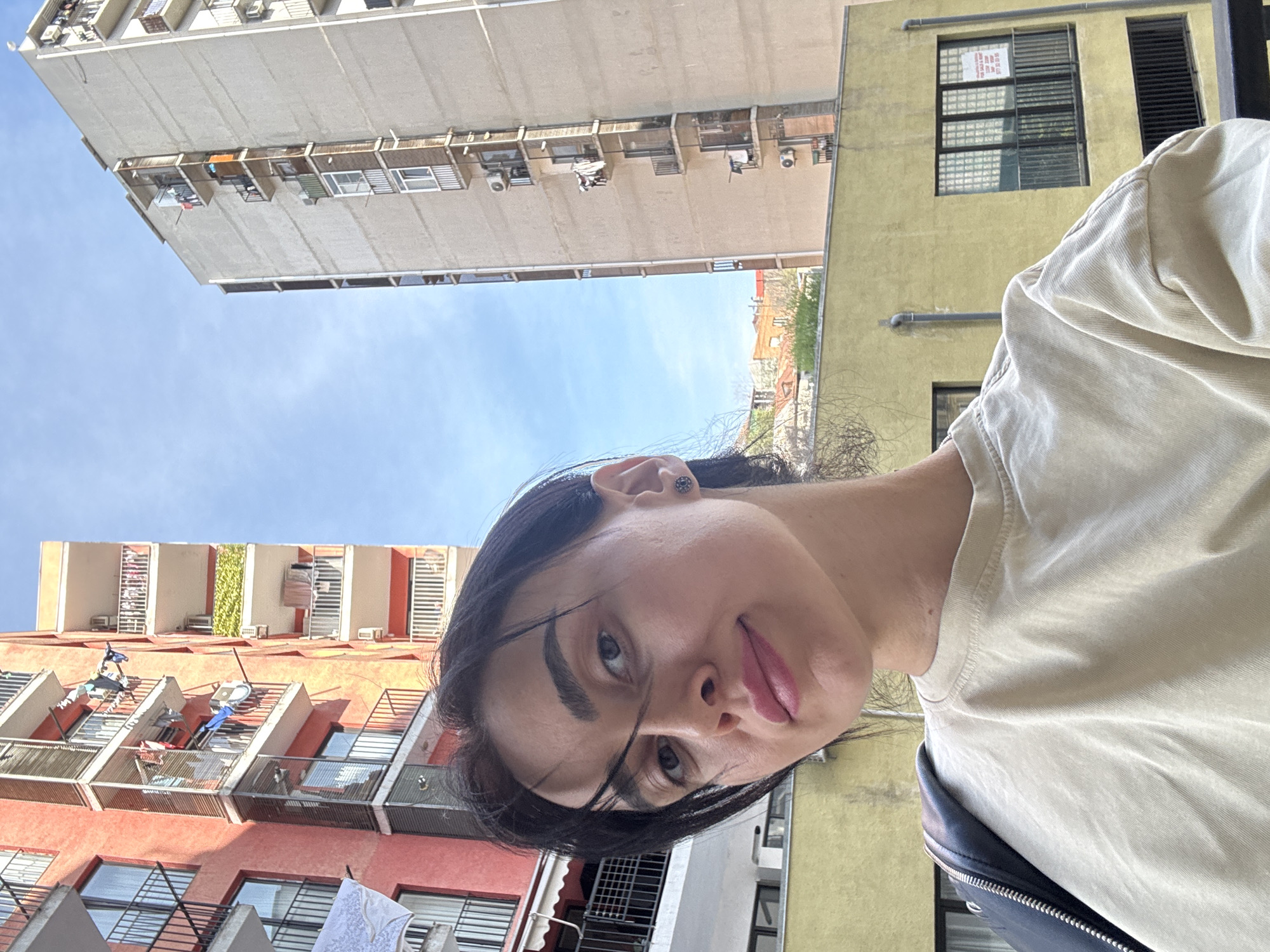 A young person with dark hair is centered in the foreground, looking down at the camera with their tongue slightly extended. The background consists of a streetscape with multiple buildings. To the left, a red building with balconies is visible, on which clothes are hanging. A tall, grey building is oriented vertically and stretches high into the frame, under a blue sky. To the right, there is a yellow building with a "for sale" sign in one of the windows. The scene appears to be an urban setting. Given the architectural style of the buildings and "for sale" sign, the location appears to be the city of Tbilisi, Georgia.