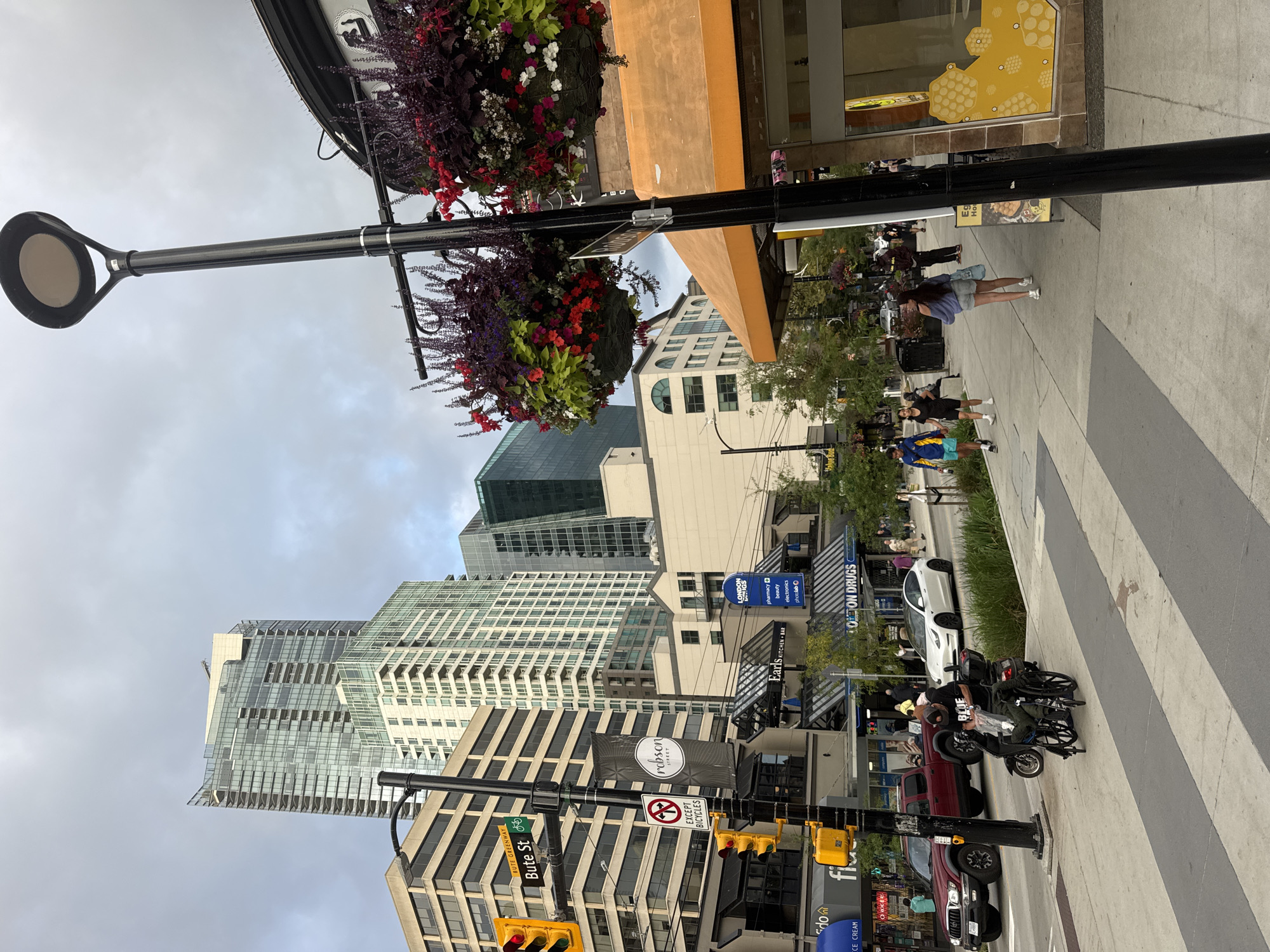 The image depicts a street scene in Vancouver, Canada, likely in the downtown area given the architecture. In the foreground, a person is seated in a wheelchair on a pedestrian crosswalk. Behind them, a red pickup truck is stopped at a traffic light. The sidewalk is lined with trees and businesses, including "Earls Kitchen + Bar" and "London Drugs." Hanging flower baskets adorn lampposts along the street. In the background, modern skyscrapers rise above the lower-rise buildings. The sky is overcast.