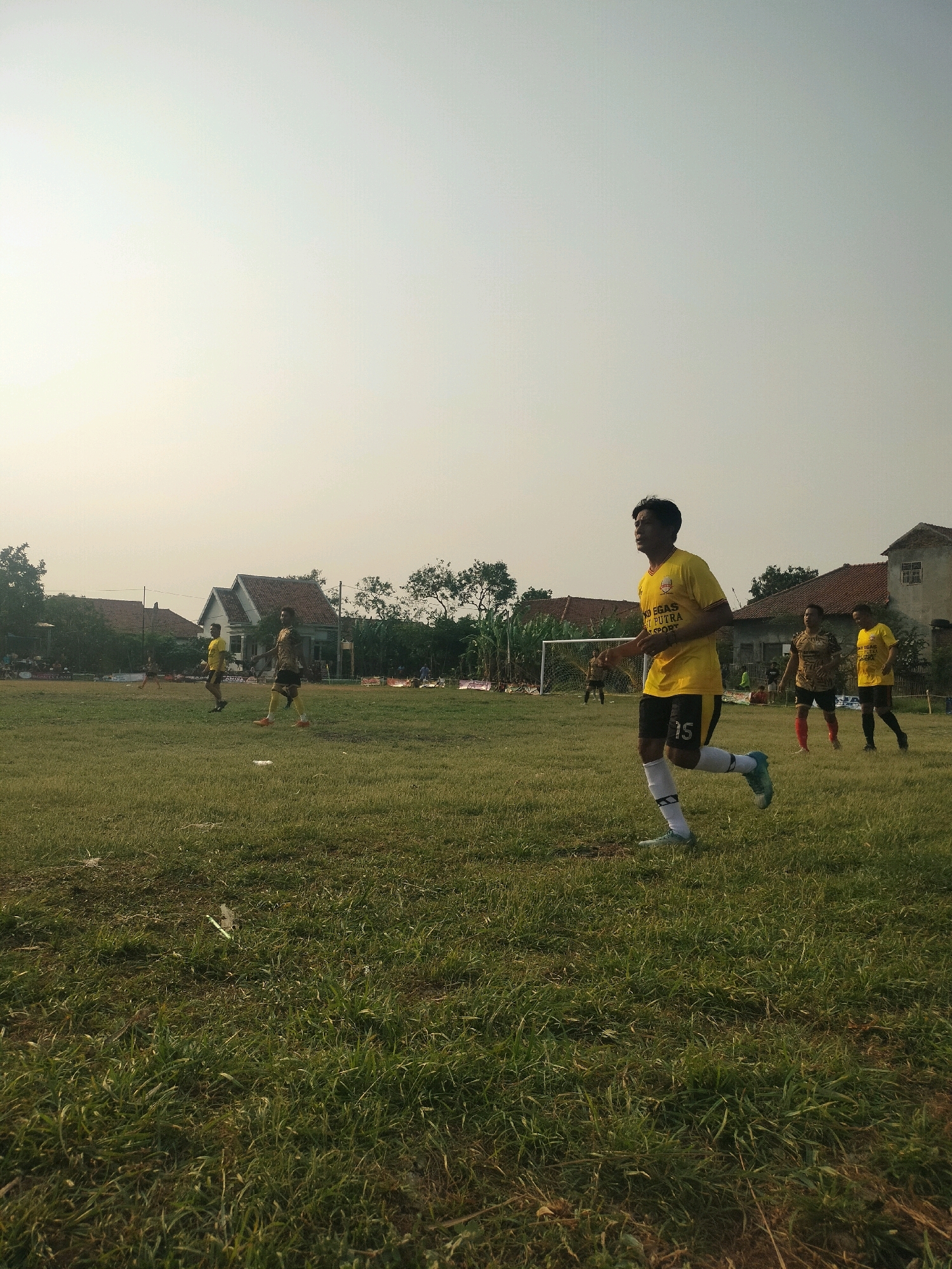 The image depicts a soccer game taking place on a grass field in Kalisari, Indonesia. The foreground is dominated by the field's green grass, with some patches of dry earth. A young man in a yellow soccer jersey with black shorts and white socks is running towards the right side of the frame, his gaze fixed ahead. The jersey has text and logos on it, with the number "15" visible on his shorts. In the mid-ground, other players in yellow and patterned jerseys are visible. A soccer goal with a white net is set up at the far end of the field. Behind the field, residential houses with red tile roofs are visible, along with trees and vegetation, suggesting a suburban or rural setting within Kalisari. The sky is a hazy light blue, indicating daylight. The overall impression is of a casual soccer match played outdoors in a local community.