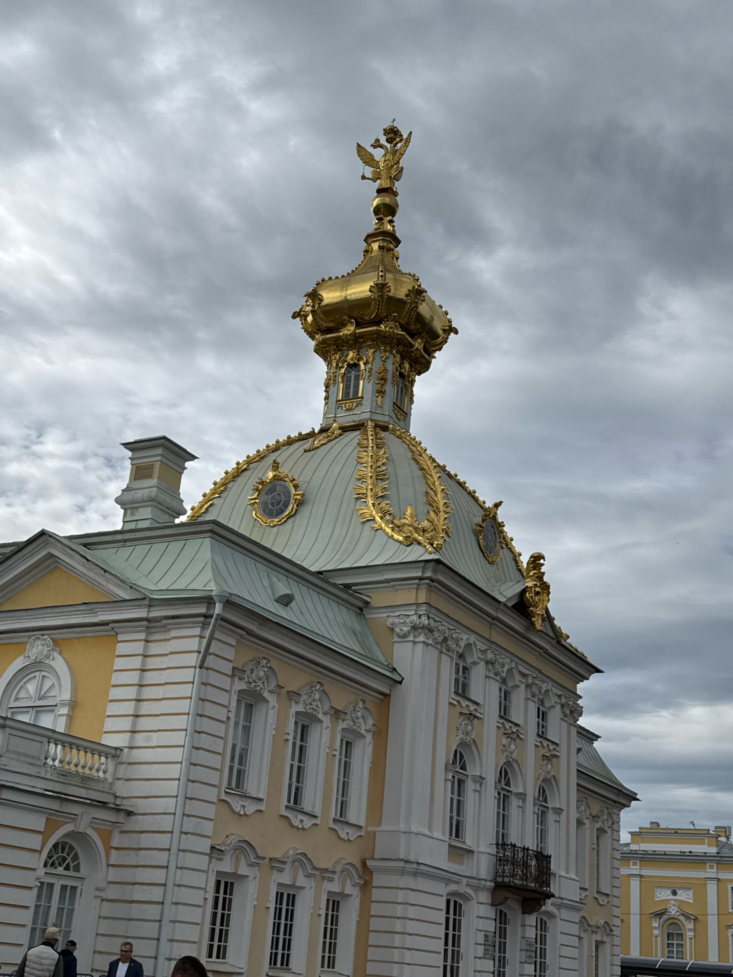 The image shows a close-up, low-angle view of the golden dome and upper facade of a historic building in Peterhof, Russia. The building is characterized by ornate golden decorations, a light green roof, and pale yellow walls with white trim. Atop the dome is a golden structure topped with a double-headed eagle emblem. The sky is overcast with heavy, grey clouds. In the lower left of the frame, a few individuals are partially visible, suggesting they are visitors or staff. Part of another yellow building with a similar architectural style is visible in the background on the right. The overall impression is of a grand, baroque-style structure under a dramatic sky.