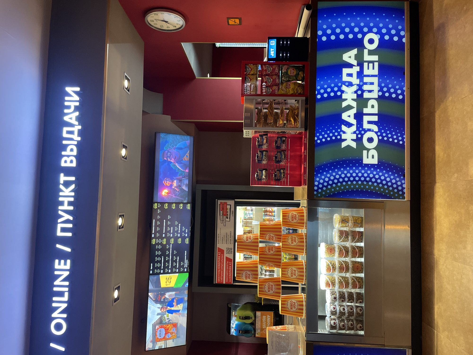 The image shows the interior of a cinema concession stand in Almaty, Kazakhstan. Above the concession stand, a neon sign reads "/ ONLINE / ПУНКТ ВЫДАЧИ". A digital screen displays movie showtimes. In front of the concession stand, there is a display of popcorn cups and various snack items such as chips and drinks. A slush machine is visible to the left of the concession stand. A clock on the wall indicates it is around 3:35. The overall environment suggests a typical cinema lobby.