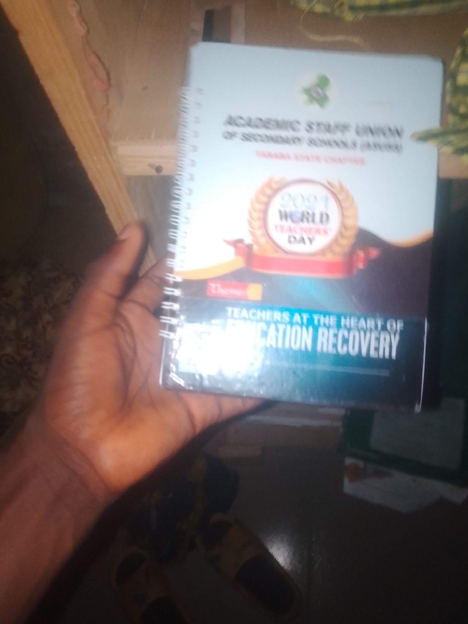 The image is a close-up, slightly angled shot of a hand holding a spiral-bound notebook. The notebook cover is light blue and features text and graphics related to the "Academic Staff Union of Secondary Schools (ASUSS)" and "World Teachers Day 2021." The theme of the event, "Teachers at the Heart of Education Recovery," is prominently displayed. The background is out of focus but appears to be an indoor environment with wooden structures and possibly some fabric. There is no discernible information to suggest the location is in Jauro Yinu, Nigeria. The hand holding the notebook is dark-skinned. The lighting is somewhat dim, with a warm tone.