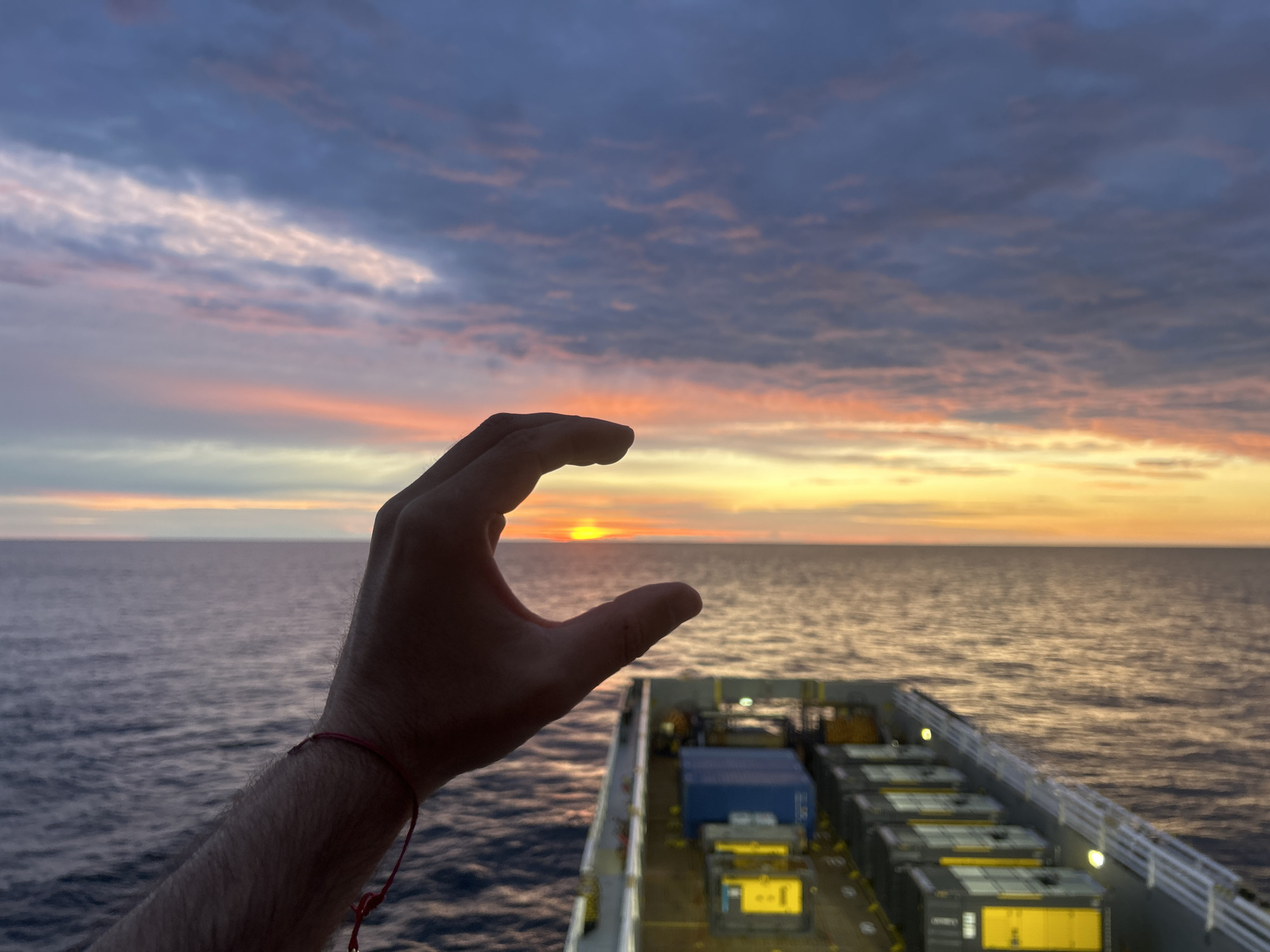 A hand, with fingers curved to form a crescent shape, is held in the foreground, obscuring a portion of the sunset. The sun, a bright orange orb, is visible through the gap between the thumb and forefinger. The hand has a red string bracelet on the wrist. In the background, the ocean stretches to the horizon, reflecting the vibrant colors of the sky, which transitions from deep blues and grays at the top to soft pinks, oranges, and yellows near the horizon. A ship, with cargo containers stacked on its deck, is positioned to the right of the frame, partially visible from a side view. The ship's deck lights are illuminated. The overall scene depicts a sunset over the ocean, viewed from the deck of a ship.