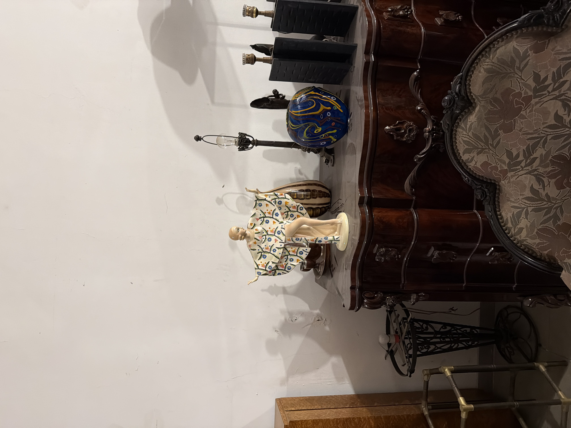 The image shows a cluttered room in Buenos Aires, Argentina. A dark wood, ornately carved dresser is placed against a white wall. On top of the dresser are several objects, including two black lamps, a blue and yellow glass ball, a lamp with a glass bulb, a vase with brown and cream stripes, and a porcelain figurine of a woman wearing a floral robe. To the right of the dresser is a portion of a high-backed chair with floral upholstery. Underneath the dresser, a vintage sewing machine base made of black wrought iron stands on the floor. Below and to the left of the sewing machine base, there is a partially visible wooden cabinet or box, and a metal structure with golden corners. The room appears to be in a state of disarray, possibly an antique store or storage space.