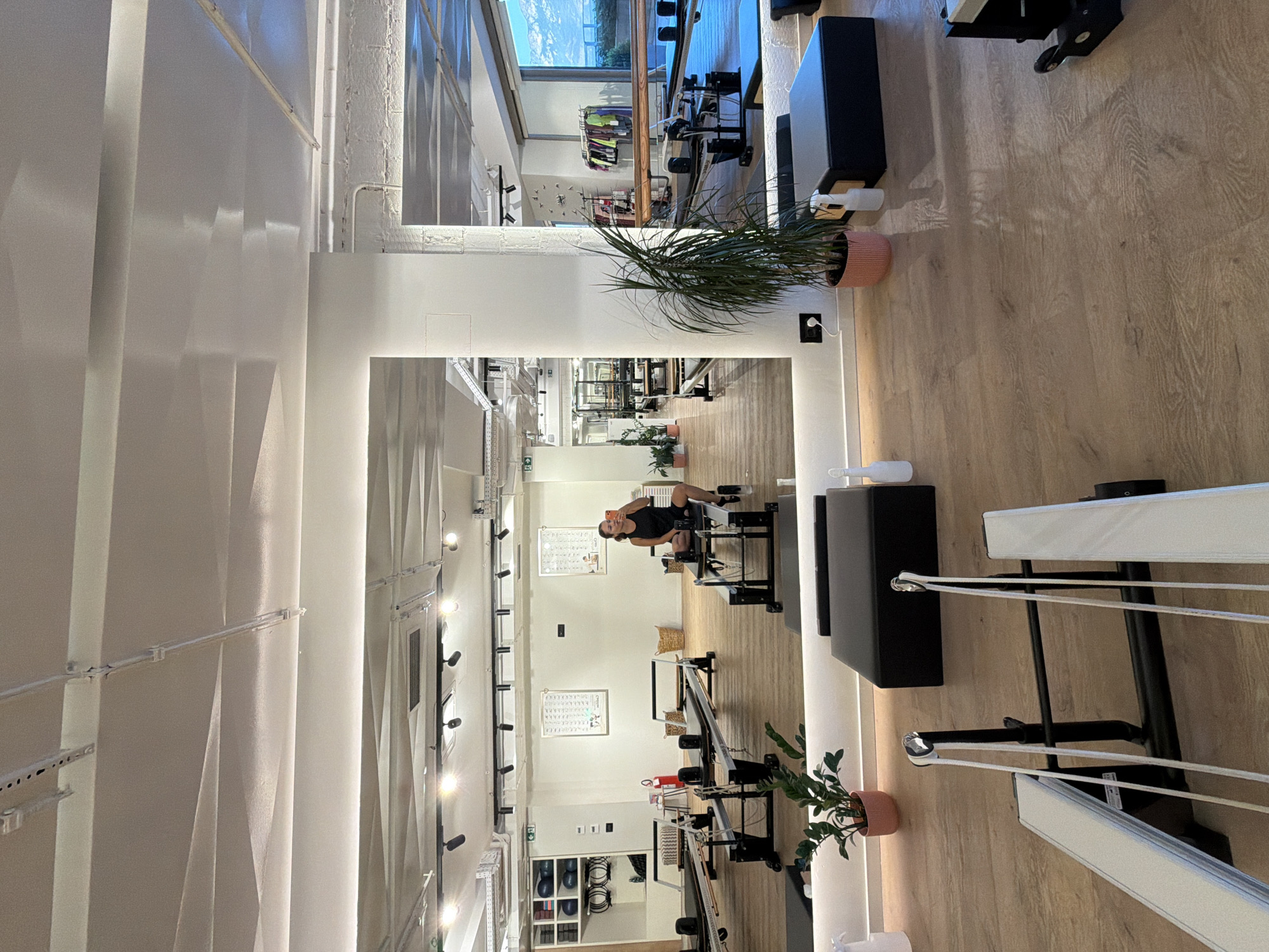 A woman is taking a selfie in a mirrored Pilates studio. The studio has wooden floors and white walls, with multiple Pilates reformers and other exercise equipment visible. There are also plants and baskets throughout the space. The woman is wearing a black tank top and shorts, and is sitting on a Pilates reformer, holding a phone in her right hand. In the background, through a large window, a mountain range is visible. The studio is located in Boreti, Montenegro.