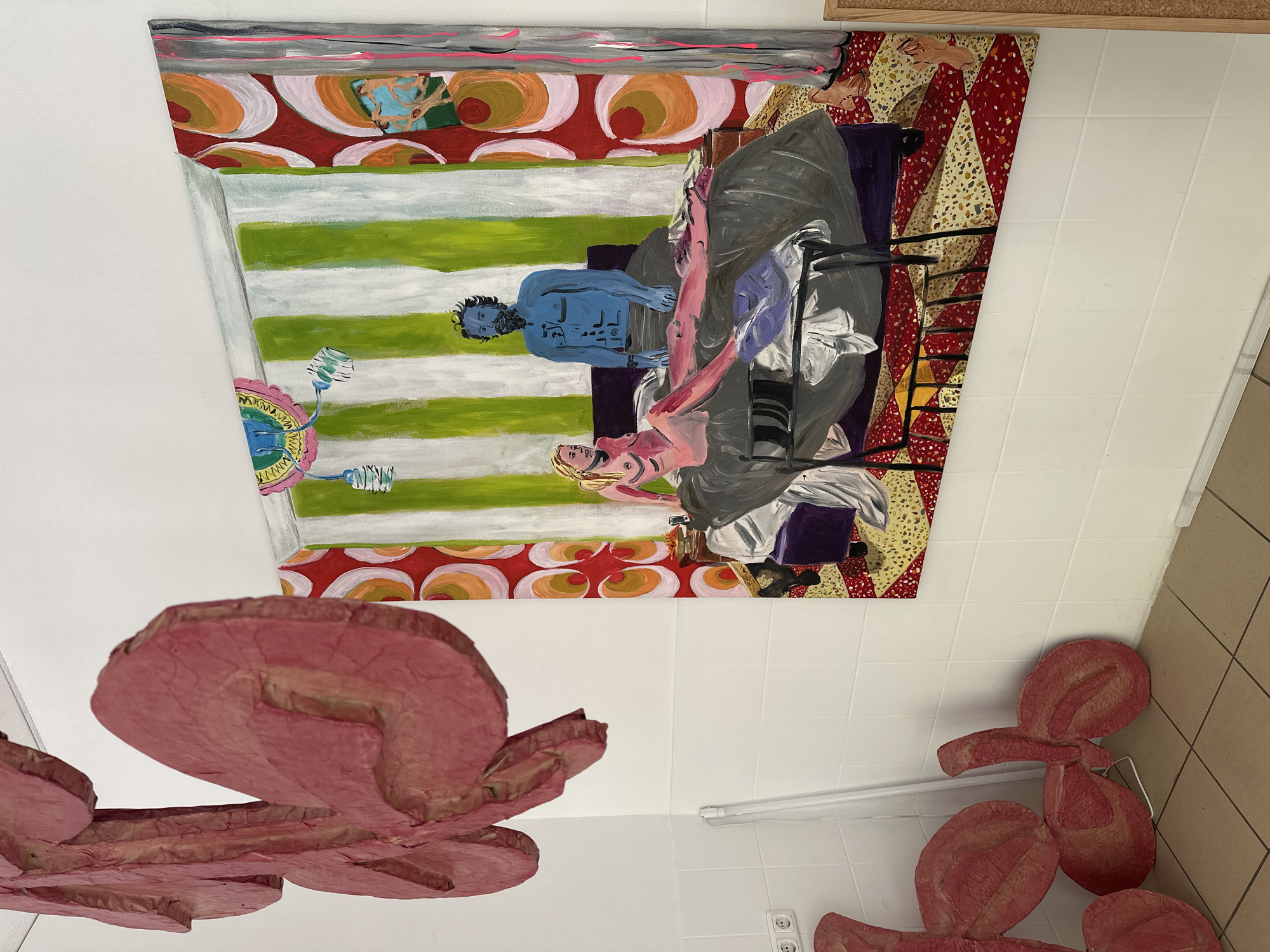 The image depicts an art installation within a room. The room has white tiled walls and a tiled floor that transitions to tan. A fluorescent light fixture is visible along the white tiled wall. On the ceiling, a painting is mounted, showcasing a scene with two figures, one painted blue and one painted pink, lying on a bed. The painting is framed with decorative borders, including geometric patterns and stripes. Below the painting are several pink, flower-like sculptures made of a textured material, possibly papier-mâché, which are affixed to the white walls and partially obstruct the view of the floor. An electrical outlet is visible near one of the flower sculptures.
Given the artistic nature of the display and the presence of tiled walls, the location could be a gallery, studio, or art space in Berlin, Germany.