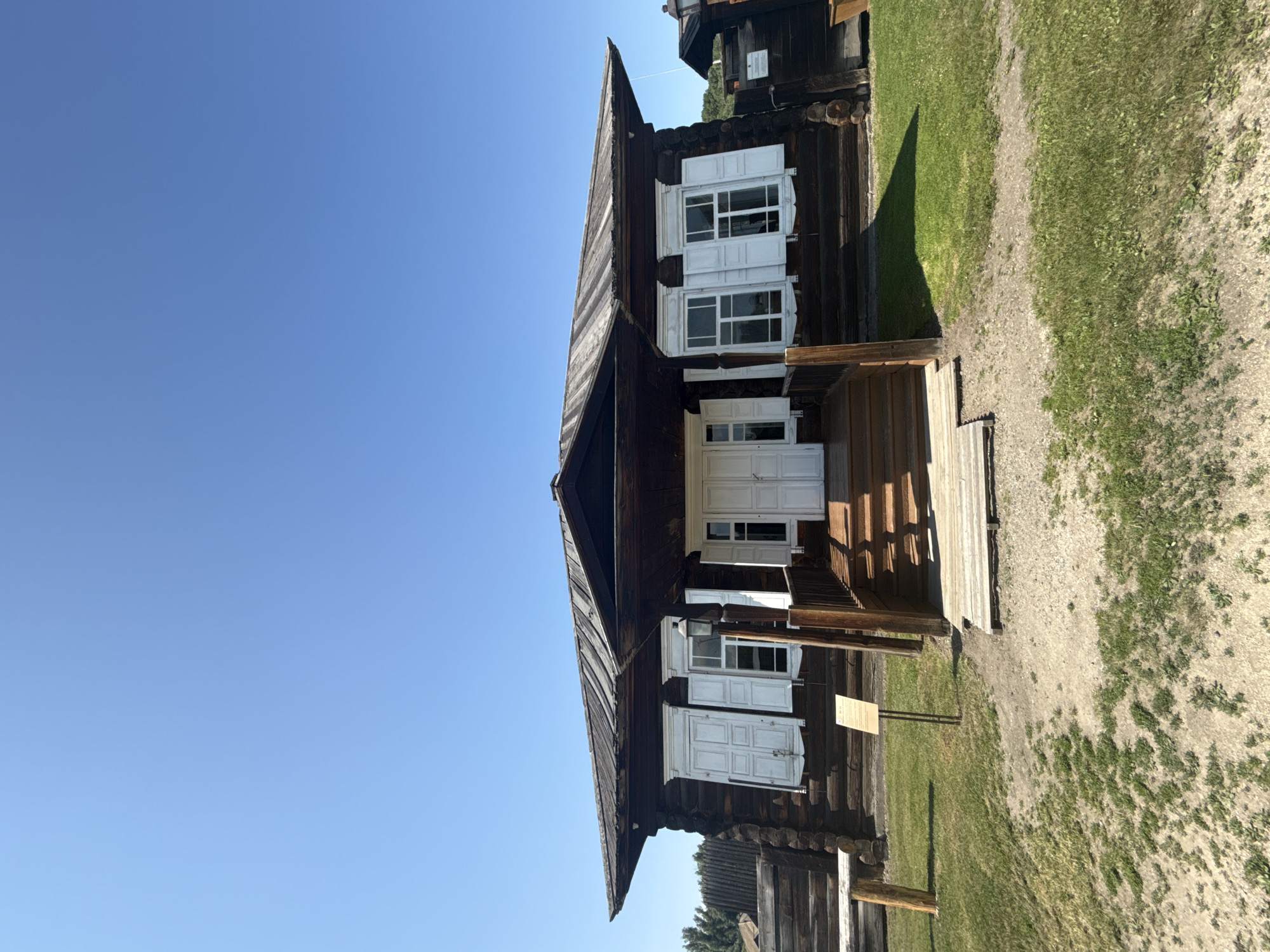 The image shows the exterior of a traditional wooden house, likely a museum exhibit, in Shushenskoye, Russia. The house has a dark wooden log construction with white shutters on the windows and white double doors leading to a porch. A wooden staircase ascends to the porch. The roof is made of weathered metal or wood. In front of the house is a grassy area with some bare patches of soil, and to the left, a wooden fence and a rustic bench are visible. A signpost stands in front of the house. The sky is clear and blue. The scene depicts a historical or cultural site, possibly related to the exile of Vladimir Lenin, as Shushenskoye is known for its Lenin memorial museum.