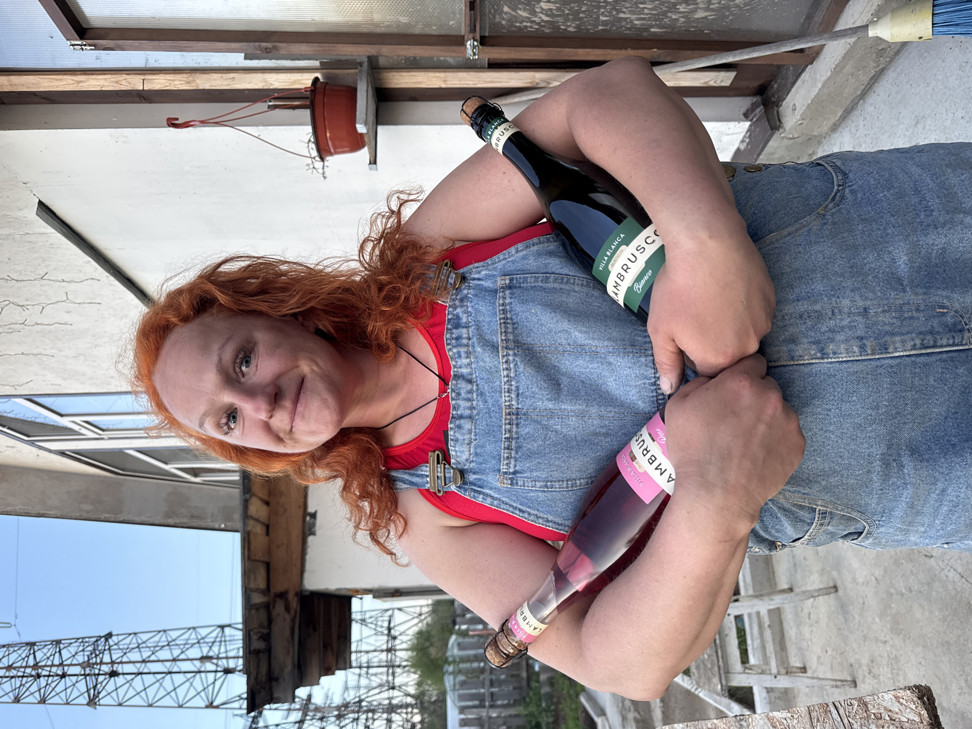 A woman with reddish-orange curly hair is holding two bottles of Villa Blanca Lambrusco, one rosé and one white. She is wearing blue denim overalls over a red top. The setting appears to be outdoors, possibly a backyard or patio, with a view of structures including scaffolding or a lattice, and what looks like an unfinished building or repairs in progress. The location is Chaikovsky, Russia.