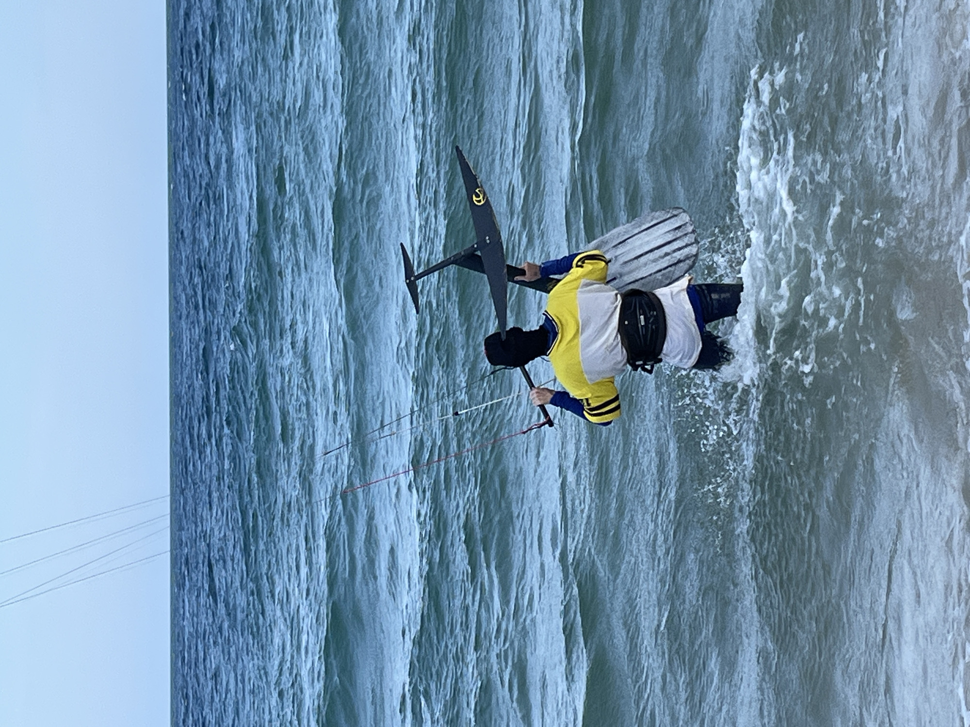 The image captures a kitesurfer engaged in the sport on a body of water. The primary subject is a person wearing a yellow and white jacket with blue sleeves and black pants, along with a black helmet. They are actively controlling a kite, which is out of frame, via a bar and lines. The kitesurfer is positioned on a hydrofoil board, a specialized board with fins that lift the rider above the water when sufficient speed is achieved.
The scene is set on a choppy body of water, likely the sea or a large lake, with visible waves and spray indicating movement. The water is a greenish-blue color, and the texture of the waves is prominent. To the left of the frame, the sky is a pale, hazy blue, suggesting either early morning or late afternoon, or possibly overcast conditions.
The kitesurfer appears to be in the midst of a maneuver, with the board slightly lifted from the water by the hydrofoil. The overall impression is one of dynamic action and outdoor recreation. There is no discernible text within the image. The lighting suggests it's daytime, and the weather appears to be windy, necessary for kitesurfing. The location cues are limited to the natural environment of a body of water, but the provided context indicates Phu Thuy Ward, Vietnam.