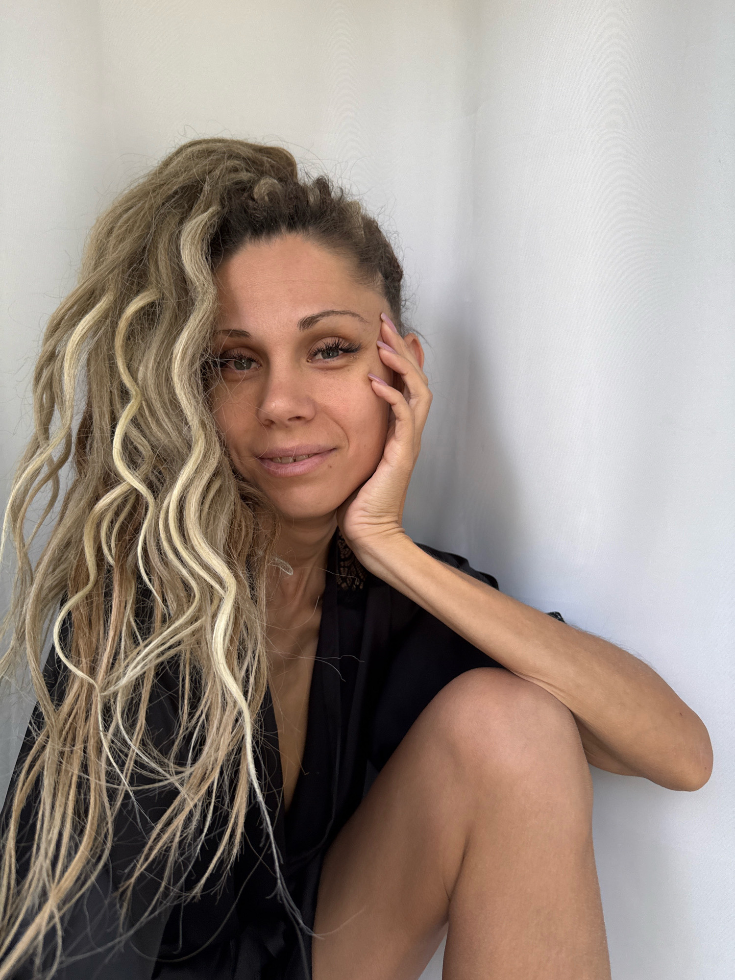 A blonde woman with dreadlocks is sitting in a corner against a white wall. She is wearing a black robe and has her hand on her cheek. The woman is looking at the camera with a slight smile. The background is a plain white wall, and the lighting is soft.