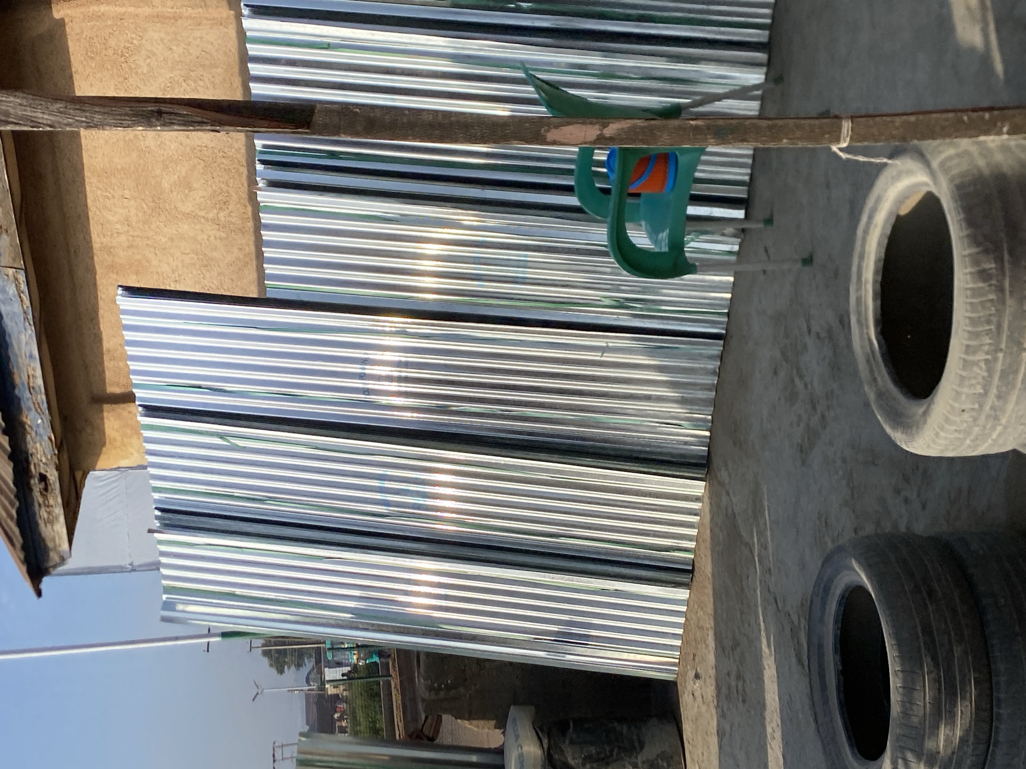 In the city of Bukuru, Nigeria, a scene unfolds outdoors with a focus on building materials. In the foreground, two large black tires are stacked on the concrete ground, suggesting a site for construction or repair. Behind them, a row of corrugated metal sheets stands vertically, leaning against a tan, textured wall of a building with a rusty, corrugated metal roof. To the right, a weathered wooden post is secured with twine. Further back, a bright green plastic chair with white legs is placed in front of the metal sheets, holding an orange and blue bucket. The background reveals a street with a few figures, some vehicles, and trees under a clear blue sky.