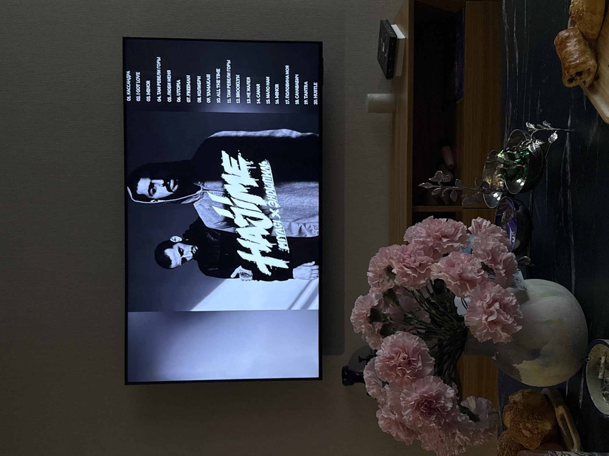 The image shows a living room setting. A flat-screen television is mounted on the wall, displaying a black and white image of two men and a tracklist. Adjacent to the television is a wooden shelving unit. A vase filled with pink carnations is placed on the table, along with other decorative objects and food.