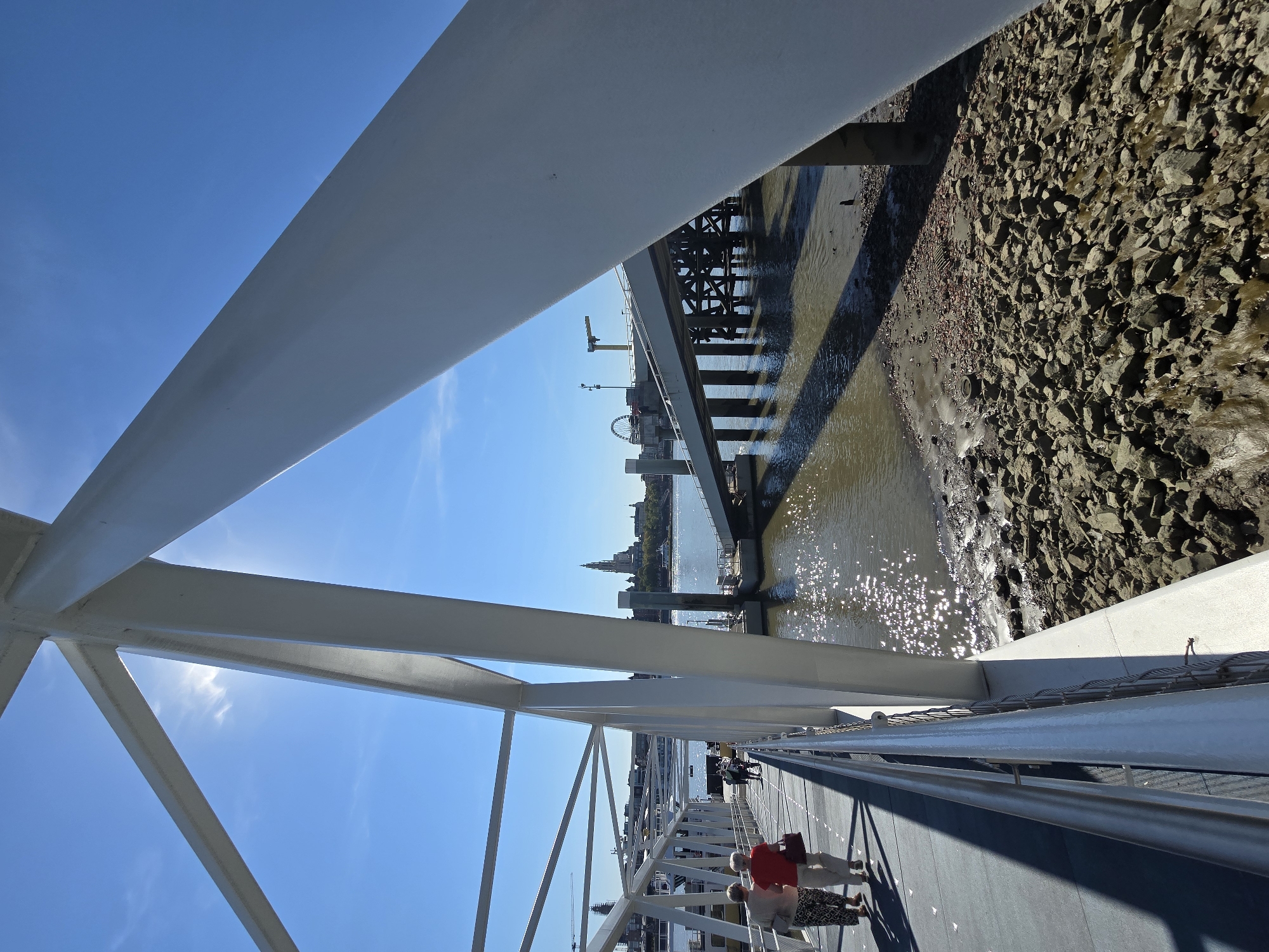 The image depicts a sunny day on the Kattendijkdok, specifically showing a pedestrian walkway leading towards a modern pier structure in Antwerp, Belgium. The walkway is made of a grey material with white railings and is bordered by large white structural supports of a bridge or pier. In the foreground, two women are walking away from the camera down the walkway. The water of the dock is visible to the right, with a rocky shore and debris along the edge. In the background, across the water, the Antwerp skyline is visible, featuring a prominent tower that appears to be the Cathedral of Our Lady, and other buildings. A Ferris wheel is also visible in the distance. The sky is clear and blue.
