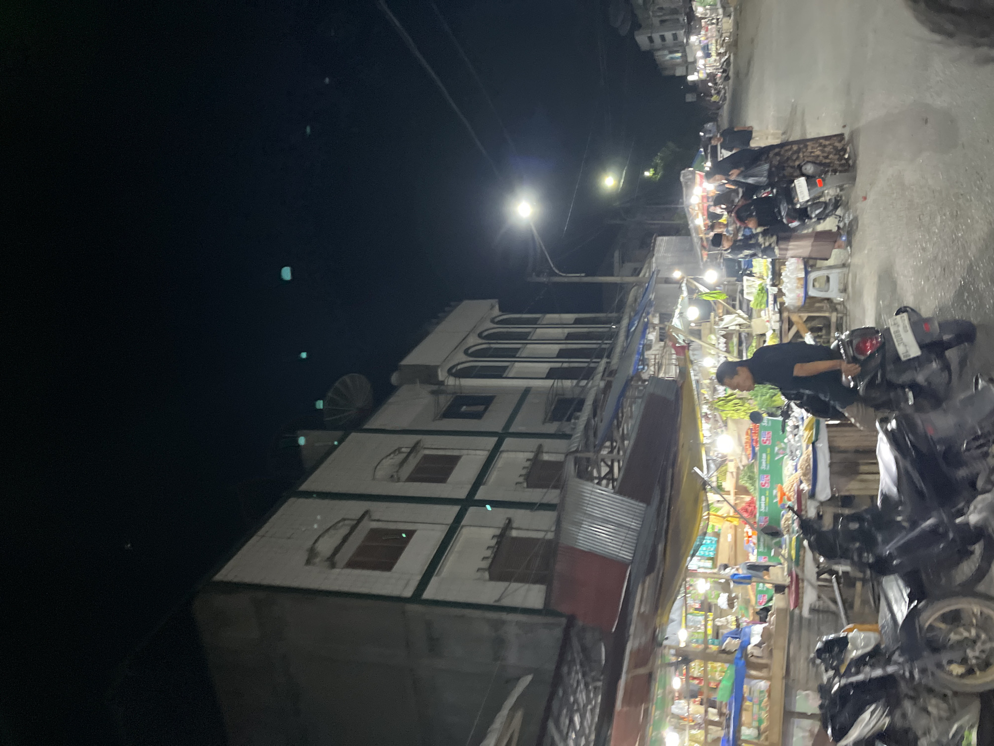 The image depicts a street scene at night in Langsa, Indonesia. On the left, a multi-story building with white walls and green trim stands prominently. Below it, a market is set up with stalls displaying various goods, illuminated by overhead lights. Several people are visible browsing or working at the market stalls. In the foreground, two motorcycles are parked. The street extends into the background, with more buildings and streetlights visible. The overall atmosphere suggests a bustling night market.