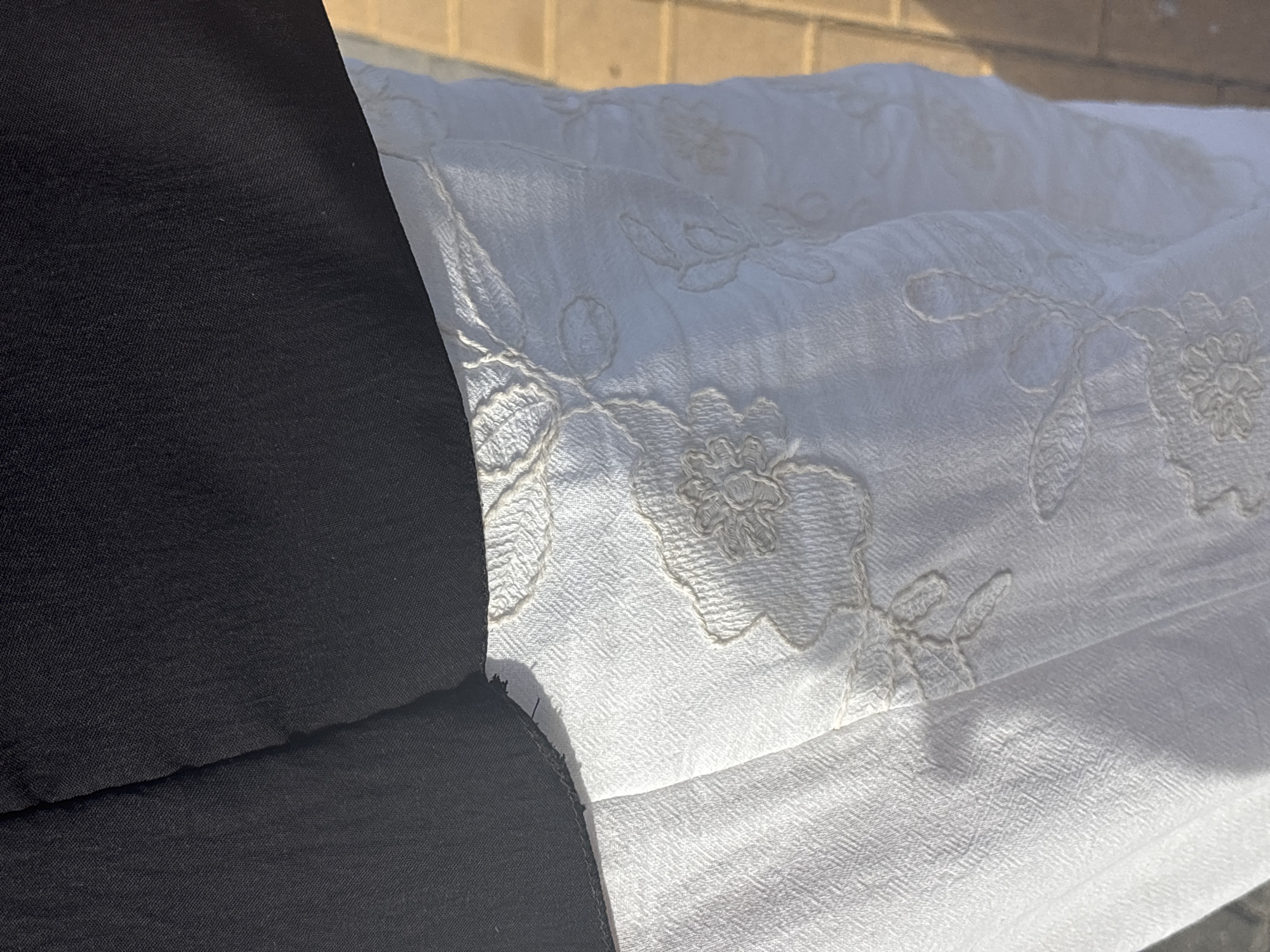 A close-up, overhead shot shows the texture of white fabric with embroidered floral patterns in a neutral tone. The fabric appears to be a skirt or dress, and is slightly creased, suggesting it is made of a linen-like material. The top portion of the image is dominated by a black garment, possibly a jacket or top, which partially obscures the white fabric. The lighting suggests an outdoor setting, with a shadow cast across the white fabric, possibly indicating sunlight. The background shows a blurred view of what appears to be a brick wall or paving stones.