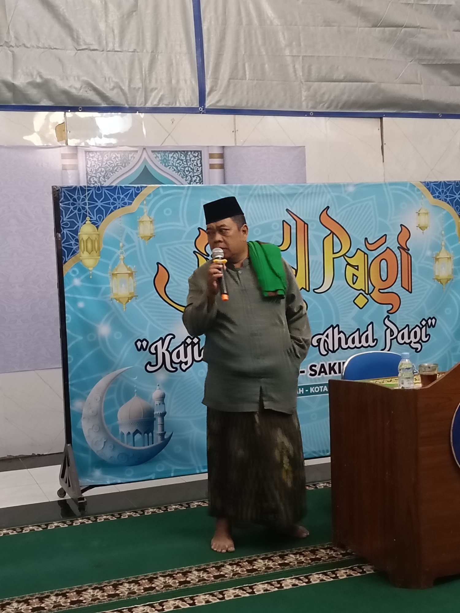 A man wearing a black peci, a green scarf, a grey shirt, and a sarong stands on a green carpet in front of a blue banner. The banner features Islamic motifs, including lanterns, a crescent moon with a mosque, and Arabic script. The man is holding a microphone and appears to be speaking. To his right, a wooden podium with a water bottle and a glass of tea is visible. The environment suggests an indoor event, possibly a religious gathering or lecture, in Kediri City, Indonesia, as indicated by the text on the banner.