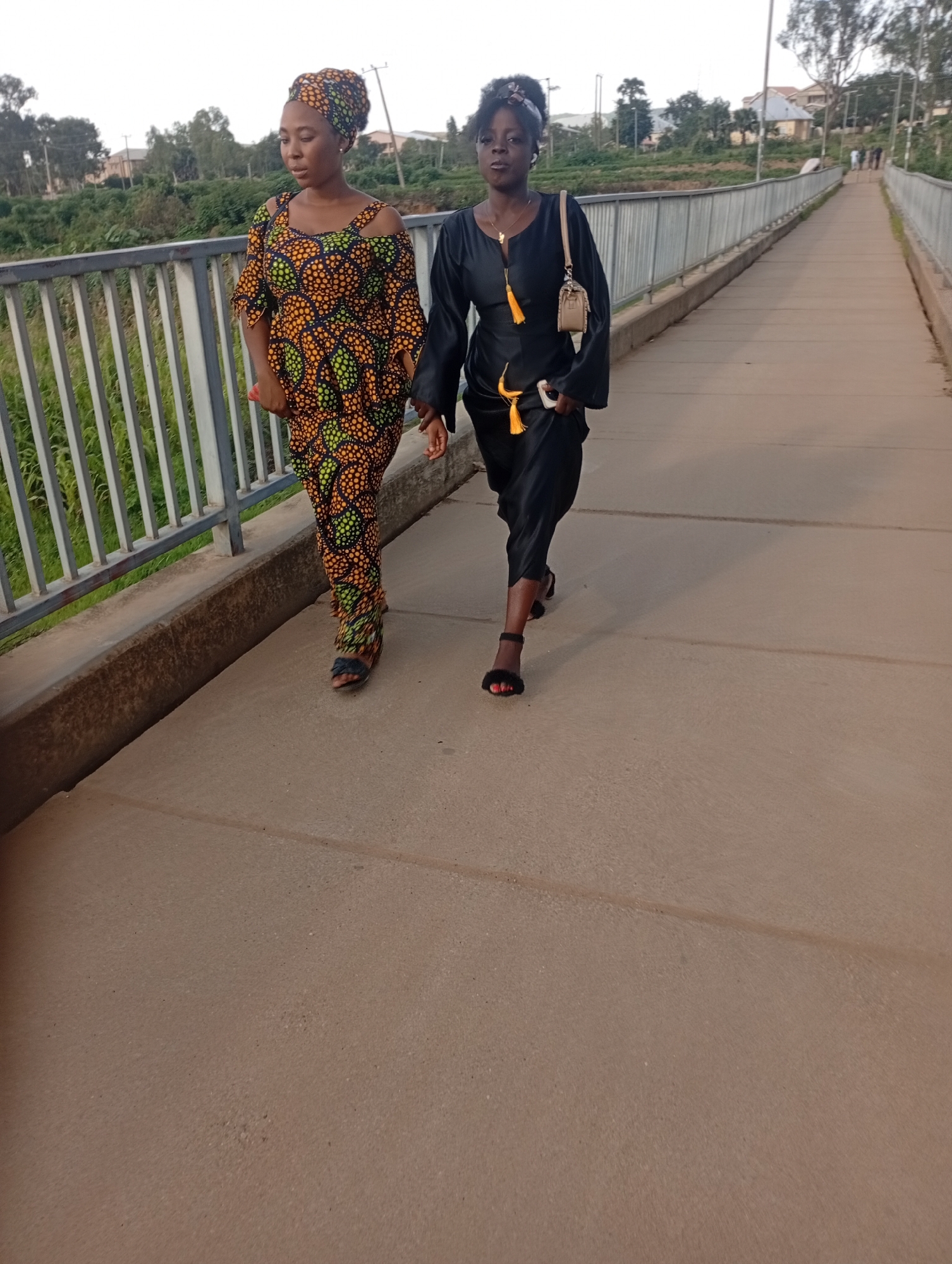 Two young women are walking on a concrete bridge in Rafin Par, Nigeria. The woman on the left is wearing a vibrant, patterned dress and headwrap, with her arms slightly bent. The woman on the right is wearing a black dress with a gold necklace and a small tan purse slung over her shoulder. Both women are facing forward and appear to be walking in the same direction. The bridge has a metal railing on the left side, and a grassy embankment with trees is visible beyond the railing. The sky is overcast.
