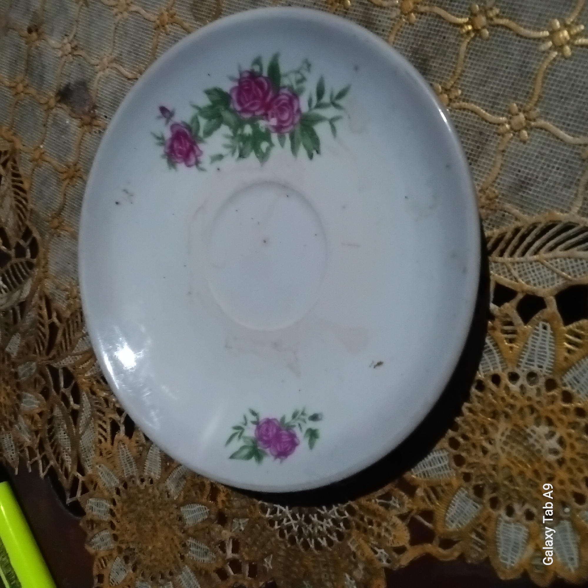 A white porcelain saucer with a floral pattern of pink roses and green leaves is centered on a decorative, golden-toned doily. The doily has a repeating diamond pattern with floral accents. A faint circular indentation is visible in the center of the saucer. The saucer is slightly dusty, with a few small dark specks on its surface. To the upper left of the saucer, a portion of a bright yellow marker is visible. The background consists of a dark surface, possibly a table, overlaid by the doily. The scene is captured from a high angle. The image is watermarked in the bottom left corner with "Galaxy Tab A9." This image was likely taken indoors in Trenggalek, Indonesia, but the exact location within the city is not determinable from the visual information provided.