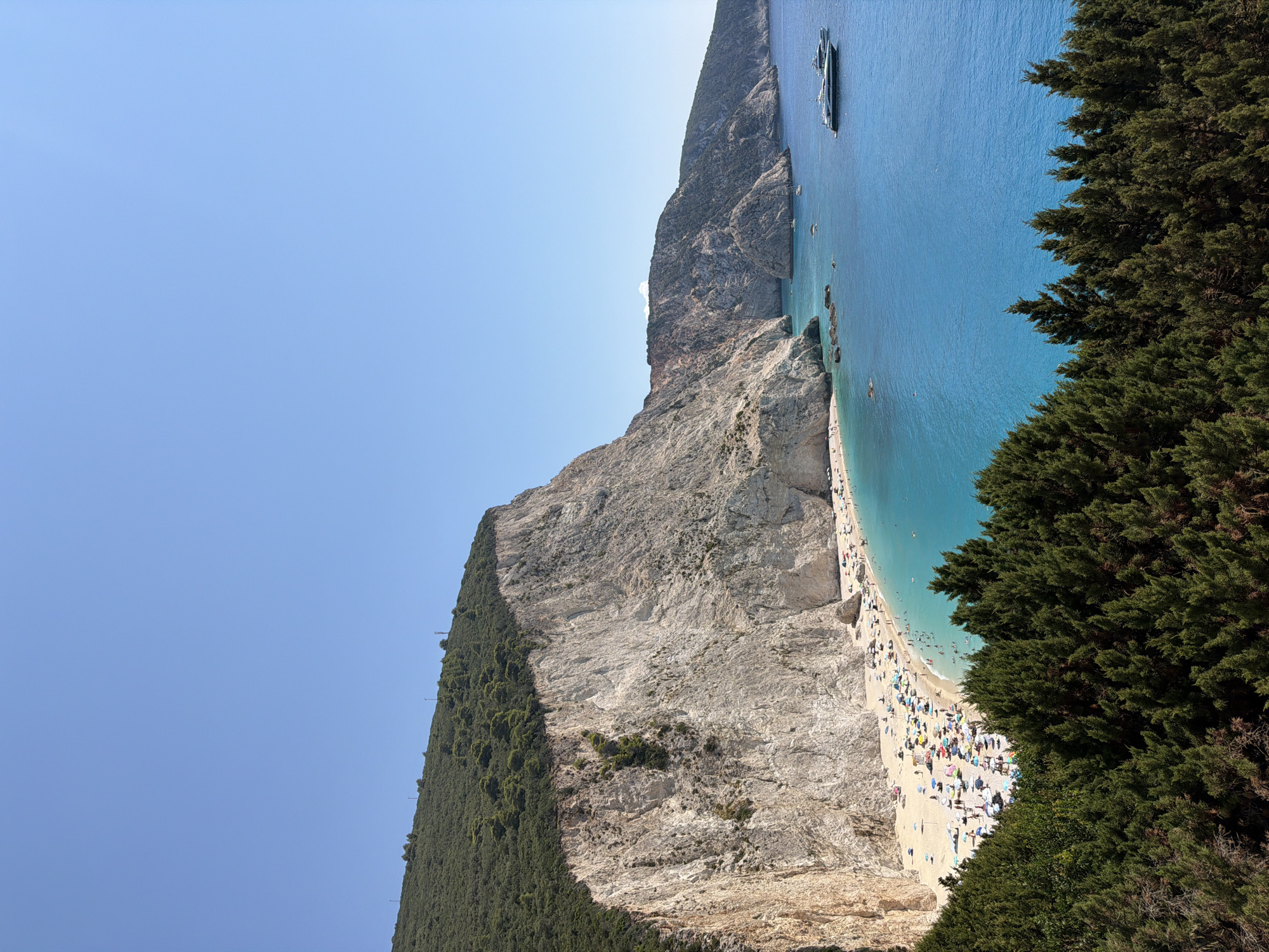 A vertical, medium shot captures a scenic view of a beach and the sea from a hillside covered in lush green trees. The pristine sandy beach is populated with numerous beachgoers enjoying the clear, turquoise waters under a bright, cloudless blue sky. A large, imposing white cliff face dramatically drops to the beach on the left side of the image, with green vegetation clinging to its upper slopes. In the distance, further cliffs and hills frame the coastline, and several yachts are anchored in the calm sea. The foreground is dominated by the dense canopy of dark green trees, creating a natural frame for the idyllic coastal scene. The location is identified as Porto Katsiki beach in the Municipal Unit of Apollonioi, Greece.