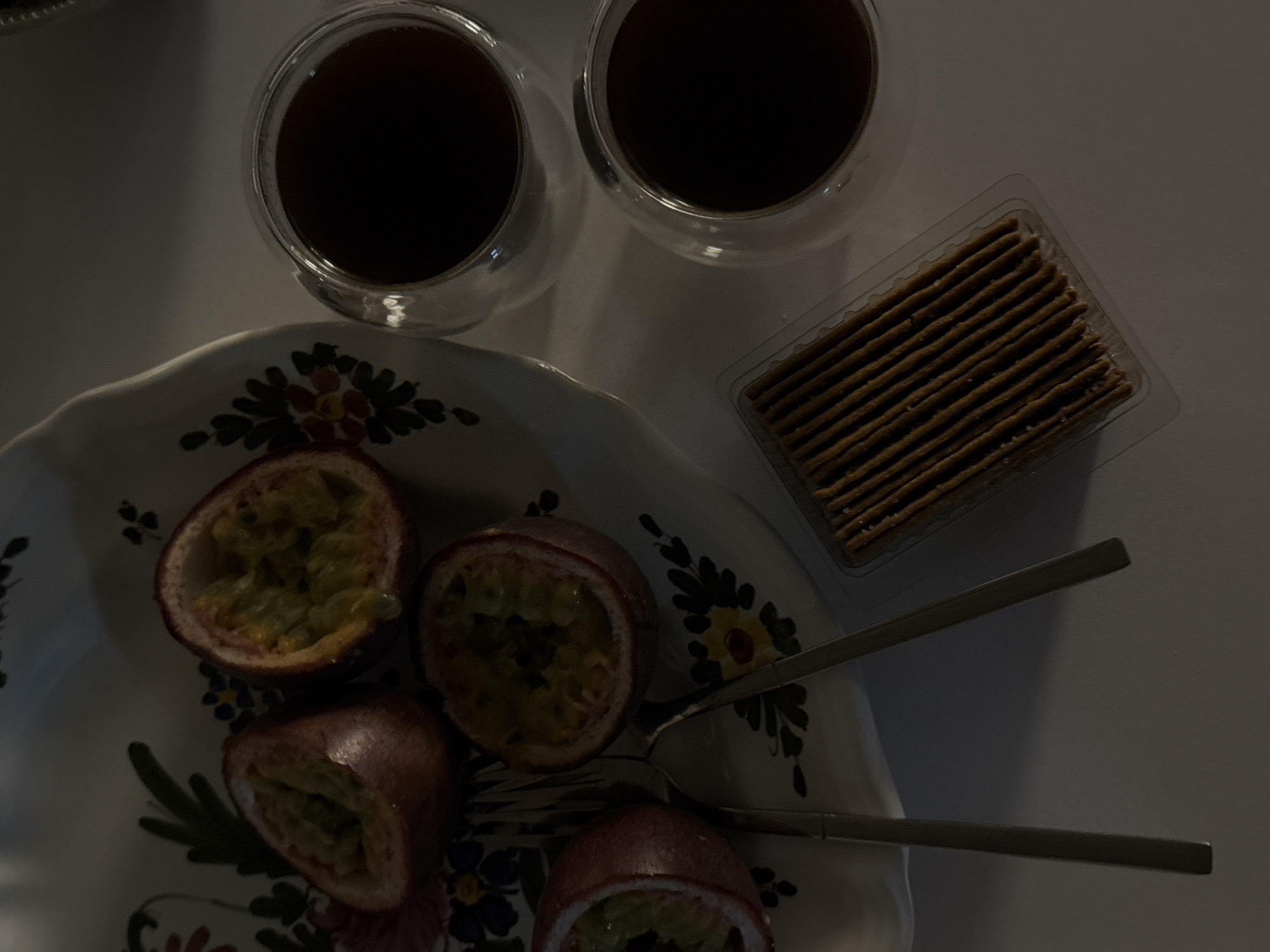 The image presents an overhead view of a table setting in Yerevan, Armenia, under dim lighting. A cream-colored, scalloped-edge plate, decorated with a floral pattern featuring dark green leaves and multi-colored blossoms, holds four halved passion fruits. The passion fruits display their internal greenish-yellow pulp and black seeds. Two silver-colored forks are positioned on the plate. To the right, two clear, double-walled glass cups contain a dark beverage, likely coffee or tea. Adjacent to the cups and below the plate, a rectangular plastic tray holds multiple rows of thin, brown rectangular biscuits or crackers. No individuals are visible, and the objects are stationary on a light-colored surface.
