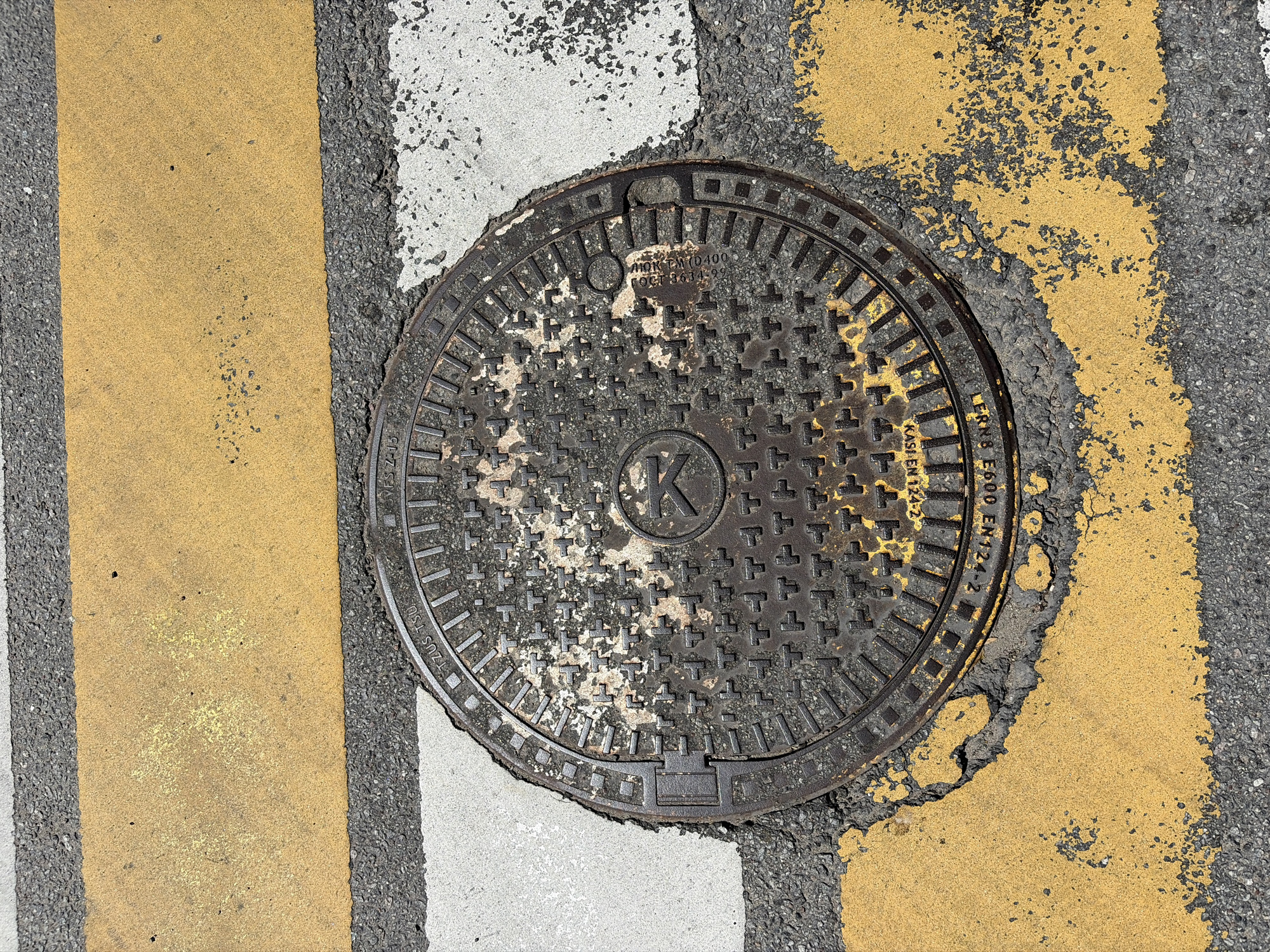 This is a high-angle, close-up photograph of a circular manhole cover set into asphalt pavement. The manhole cover is dark gray and heavily textured with a repeating pattern of cross-like shapes and a large letter "K" in a circle at its center. There are some areas of wear and possibly rust on the cover, with traces of yellow paint also visible.
The manhole cover is surrounded by asphalt, which is marked with sections of road paint. To the left of the manhole cover, there is a thick, bright yellow stripe, bordered by a thinner white stripe. To the right and partially obscuring the manhole cover, there is a larger, irregularly shaped patch of yellow road paint. The asphalt itself is dark gray and appears slightly rough.
The lighting suggests it is daytime, possibly a sunny day, given the brightness and the way the light interacts with the textured surfaces. There are no people or other living beings visible in the image. The focus is entirely on the manhole cover and the surrounding road markings.
Visible text on the manhole cover includes "TZUS TSU", "ΛΙΟΝ ΤΜ (D400", "ГОСТ 3634-99", and "LERNS E600 EN124-2...". The text "KASI EN 124-2" is also visible. This text likely indicates the manufacturer, model, load rating (D400 suggests a load capacity of 400 kN), and relevant standards for the manhole cover. The context suggests this is an outdoor scene, likely a street or pedestrian area where such manhole covers are common.
