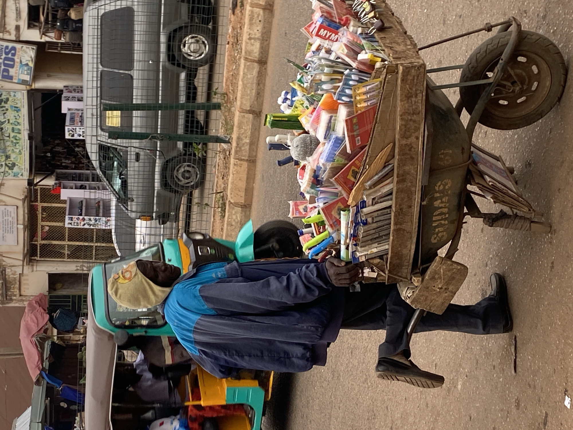 This photograph captures a street vendor pushing a loaded cart in Bukuru, Nigeria, likely during the daytime under an overcast sky. The image is taken from a slightly elevated perspective, looking down at the vendor and his merchandise.
The primary subject is a man, presumably the vendor, dressed in a blue and black long-sleeved jacket and dark trousers, with a yellow cloth wrapped around his head. He is positioned behind a large, two-wheeled cart that he is pushing along a paved surface. The cart is overflowing with various items, predominantly stationery and small household goods, including pens, pencils, notebooks, toothbrushes, and other packaged products. Several items have visible brand names, such as "MYM," "BESK," and "STAR." The cart itself appears to be a repurposed or modified wheelbarrow, painted olive green, with the letters "SDAS" or "SIDAS" partially visible on its side. The vendor wears dark loafers and appears focused on his task.
In the background, to the left, a grey van is parked behind a metal fence, suggesting a commercial or residential area. Further back, a vibrant green and yellow tricycle, commonly known as a Keke Napep in Nigeria, is visible, possibly parked or moving slowly. Another person, likely the driver, is partially visible within the tricycle. The background also features buildings and shops, with one sign clearly readable as "POWER POINT POS SERVICE HERE." The overall scene depicts a bustling, everyday street environment characteristic of urban or semi-urban areas in Nigeria.
