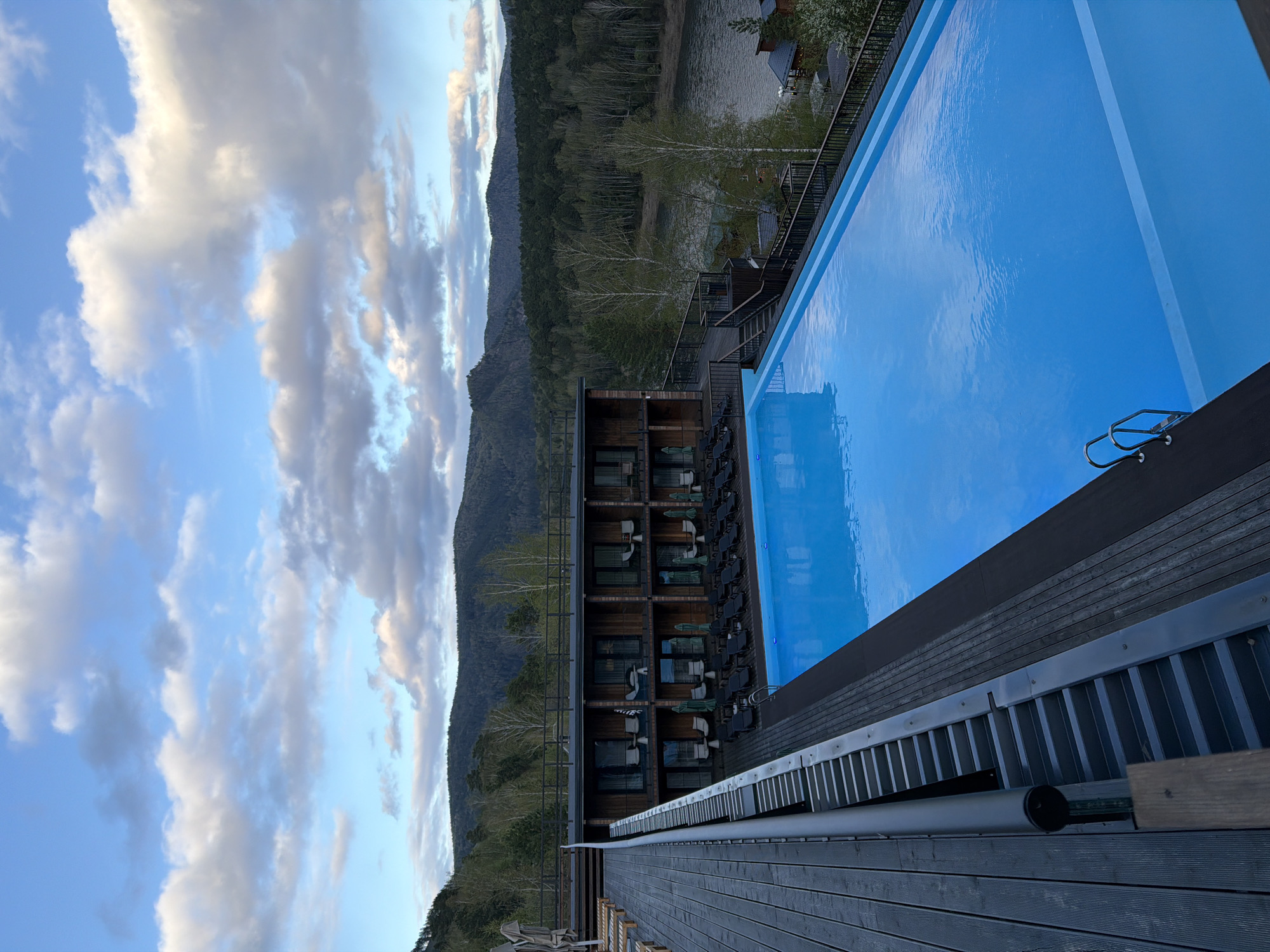 The photograph presents an outdoor scene featuring a rectangular swimming pool, a multi-story building, and a natural landscape. The pool's water reflects the sky, creating a vibrant blue surface, with a ladder visible at one end. Adjacent to the pool is a wooden deck. The building exhibits a modern architectural style, with balconies and windows. In the background, a range of mountains and a body of water can be seen under a cloudy sky. It is located in the city of Манский, Russia.