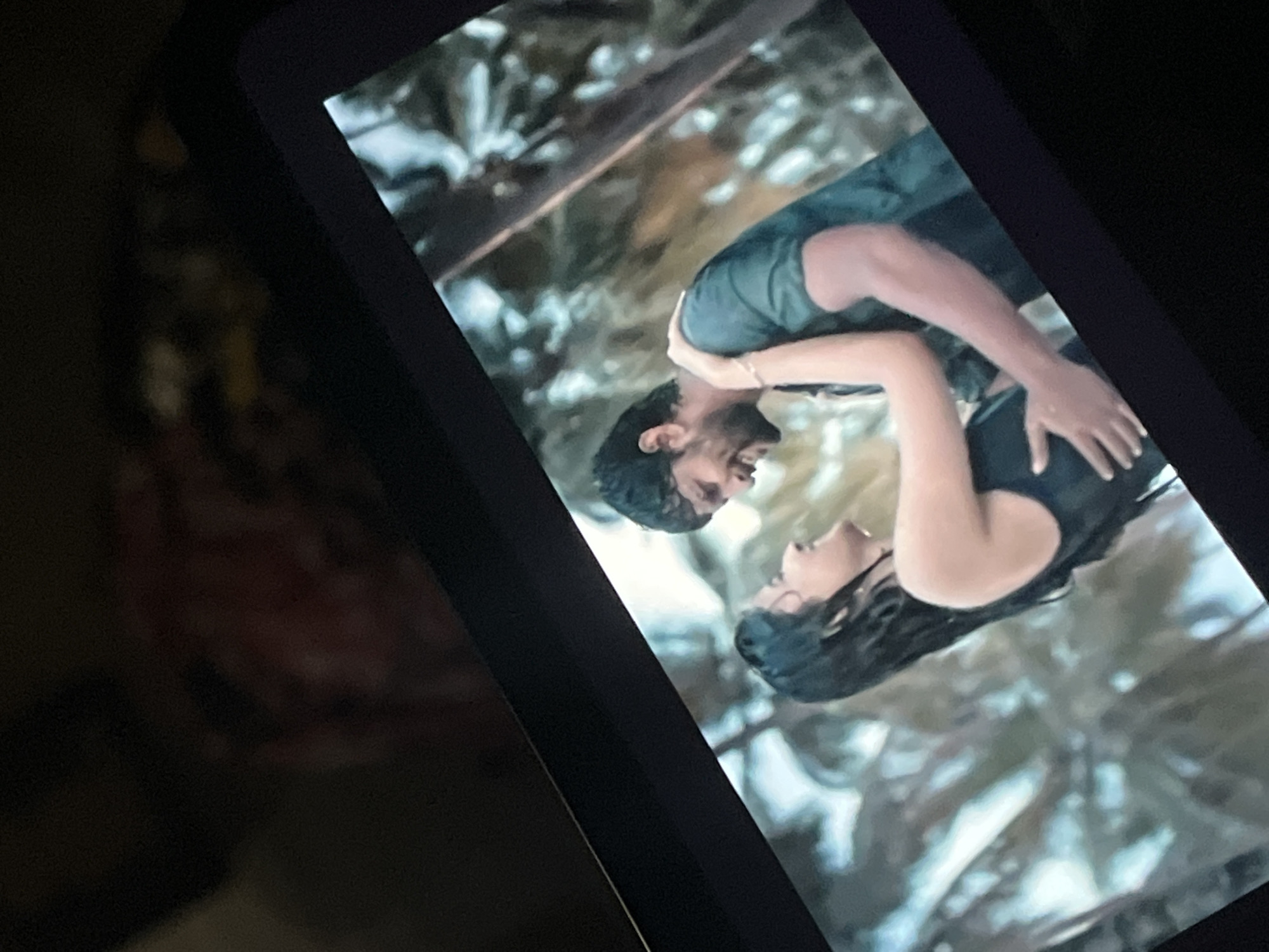 The image shows a close-up of a tablet screen displaying a movie scene. The scene features a man and a woman embracing outdoors. The man has dark hair and is wearing a dark green t-shirt. The woman has long, wet dark hair and is wearing a black sleeveless top. They are both smiling and appear to be in a playful or romantic interaction, possibly in the rain or near water, with lush green foliage in the background. The tablet is angled, and the surrounding environment is dark, suggesting it is being used indoors at night. No specific location within Bukuru, Nigeria can be identified from the image content.