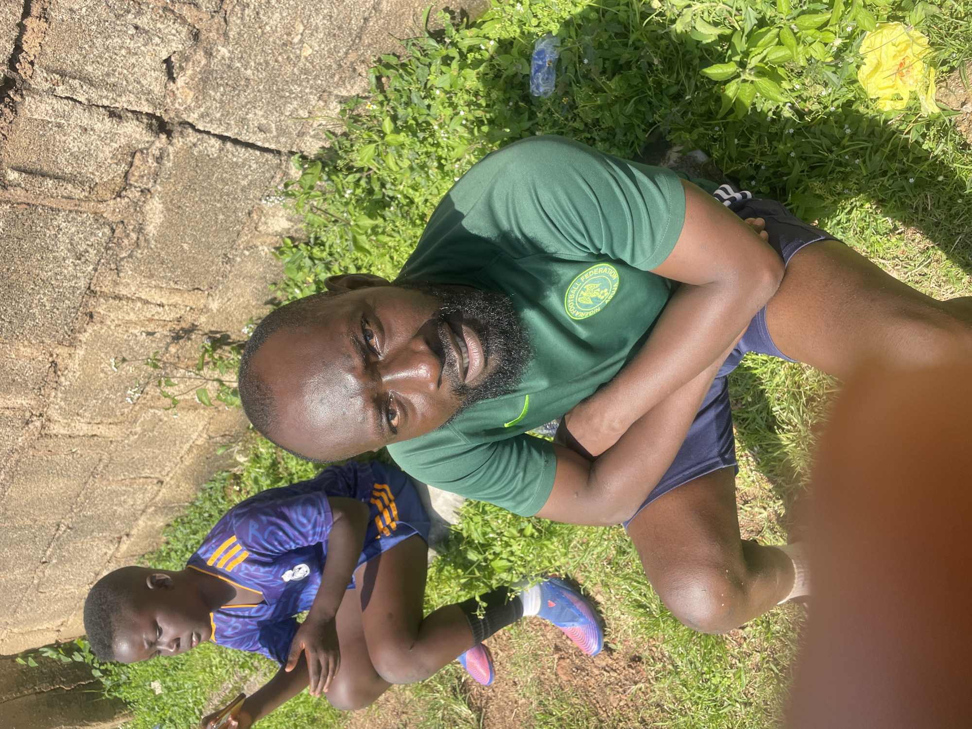 Two individuals, one adult male and one child, are sitting on the ground near a rough-hewn stone wall in a grassy area. The adult male, positioned in the foreground and facing the camera, wears a green t-shirt with a circular logo on the left chest, dark shorts, and has a beard. The child, in the background and looking to the left, is wearing a purple and orange soccer jersey and dark shorts. The environment appears to be outdoors, with green grass and a textured stone wall providing a backdrop. A yellow flower is visible in the lower right corner of the image. The lighting suggests a sunny day. The overall scene depicts a casual outdoor moment, potentially during or after a sporting activity, given the attire of both individuals.