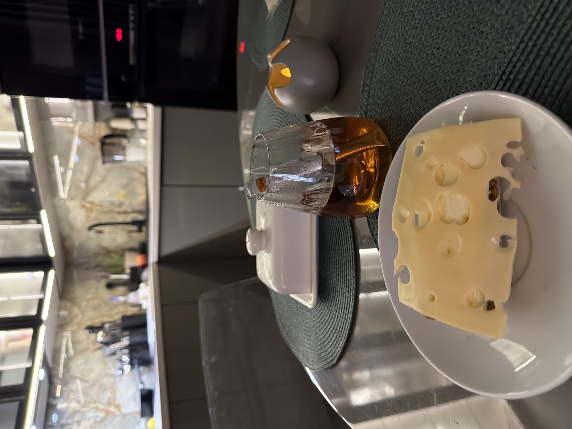 The image shows a close-up of a slice of Swiss cheese on a white plate, next to a glass of amber-colored liquid. A small, decorative candle holder with a lit candle is also visible. The scene appears to be set on a kitchen counter, with a blurred background that suggests a modern kitchen with marble countertops and stainless steel appliances. The lighting suggests it might be evening or indoors with artificial light. There is no visible text. The overall mood is calm and relaxed, possibly indicating a moment of personal indulgence or a quiet evening at home. The cheese has distinctive holes, typical of Swiss varieties. The drink in the glass is likely tea, whiskey, or brandy. The plate is resting on a dark green placemat.