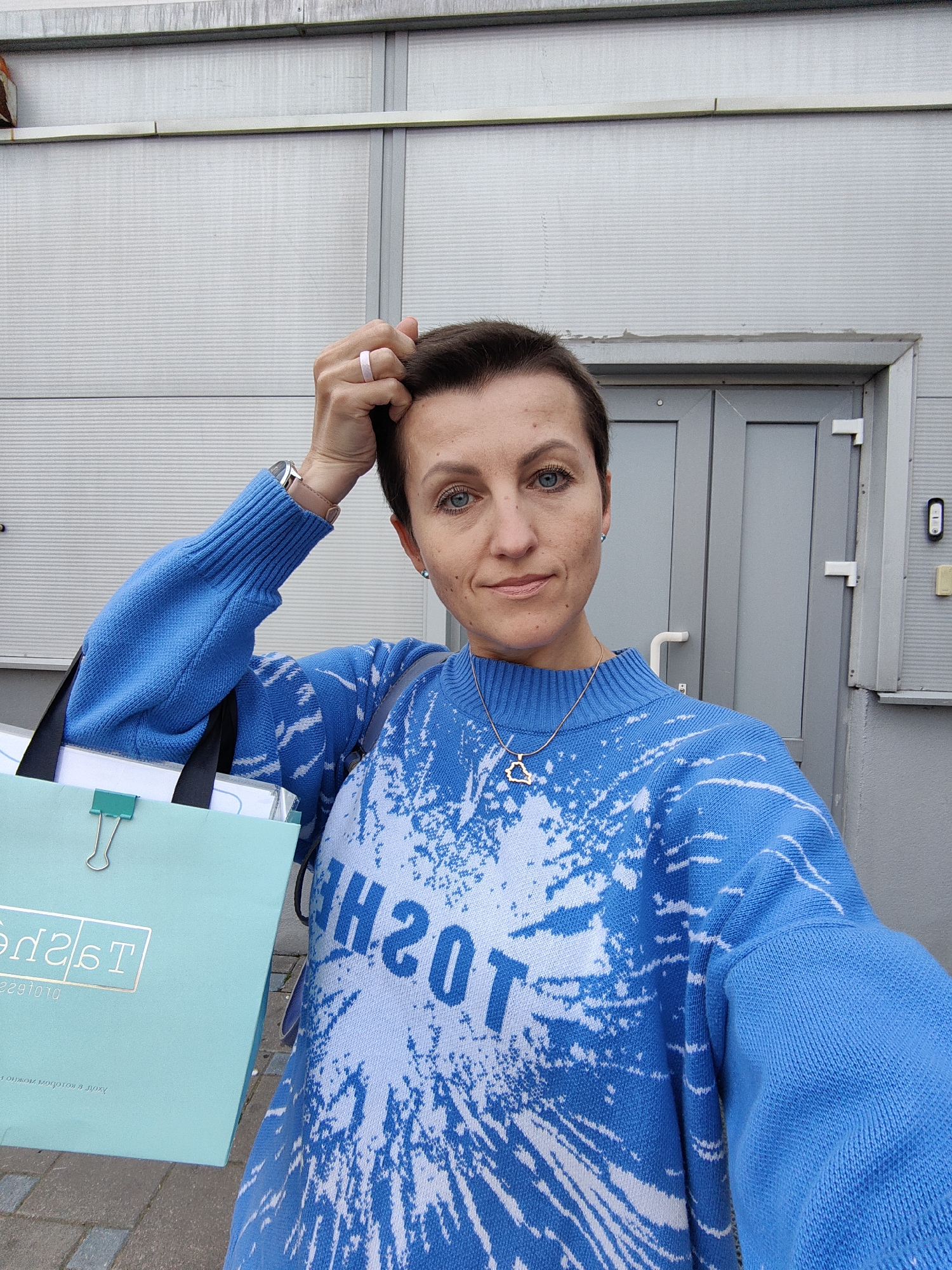A woman with short brown hair and blue eyes, wearing a blue sweater with white abstract patterns and the word "TOSH" printed on it, stands in front of a gray building with two doors and a metal facade. She is holding a light blue shopping bag with the text "Tad's" visible and a green binder clip. A silver wristwatch and a pink ring are visible on her left wrist and finger, respectively. A delicate silver necklace with a pendant is worn around her neck. The image was taken outdoors, likely in the city of Minsk, Belarus, given the context provided. The background consists of a gray building and a paved walkway. The lighting suggests it is daytime.