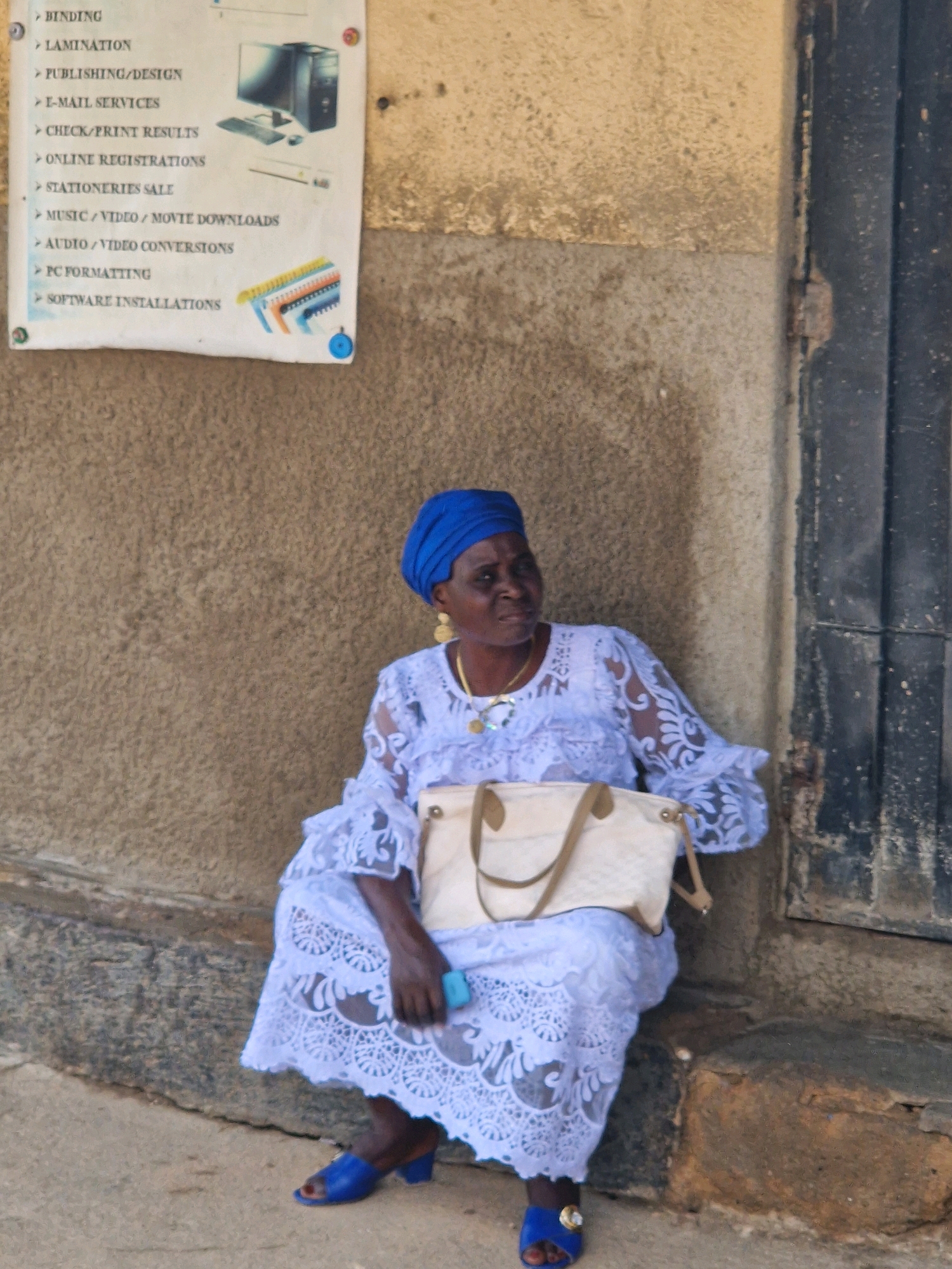 A full-shot, eye-level, outdoor photograph captures an older Black woman seated on a step against a textured, beige concrete wall. She is wearing a bright blue headscarf, a white lacy dress, and blue open-toed heels. The woman is holding a light-colored handbag on her lap and a small blue object, possibly a phone, in her right hand. A sign with text and an illustration of a computer is affixed to the wall behind her. The scene appears to be in an urban environment, with the presence of a dark metal door frame to the right of the woman. The lighting suggests it is daytime, with soft shadows cast by the woman and the wall.