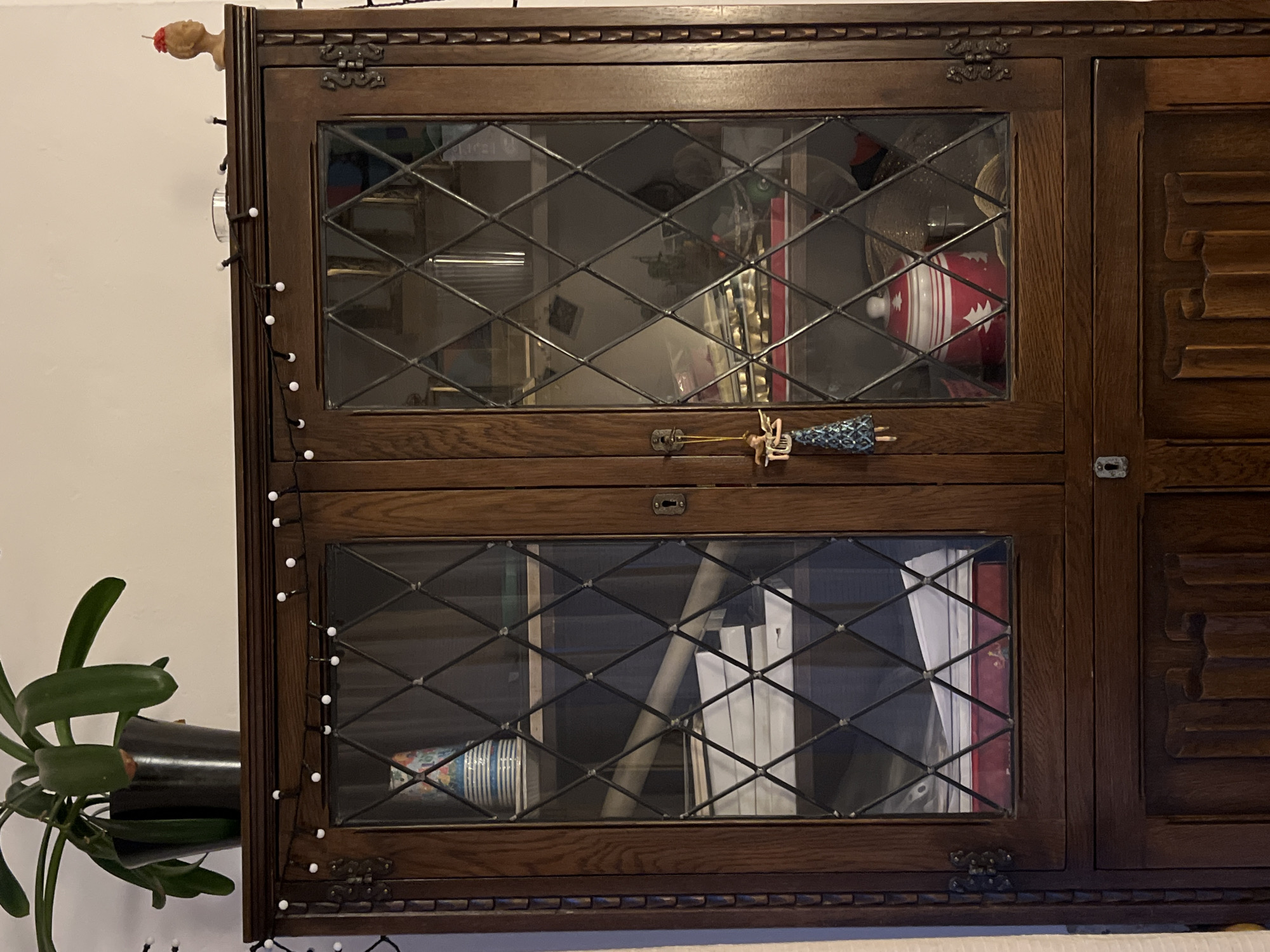 An oak display cabinet stands against a light-colored wall. A potted plant with large green leaves sits on top of the cabinet, to the left. The cabinet has two glass-paned doors with diamond-shaped mullions, each with a decorative keyhole and handle. A small, festive ornament, possibly an angel, hangs from the handle of the left door. The shelves inside the cabinet are filled with various items, including stacks of papers, books, and decorative containers. A string of small white lights is draped across the top of the cabinet. On the right side of the cabinet's top, a small, carved wooden bust with a red object on its head is visible, along with a small glass votive holder. The bottom section of the cabinet consists of two wooden doors with carved detailing. The overall impression is one of a domestic interior, likely a living room or dining room, in Sofia, Bulgaria.