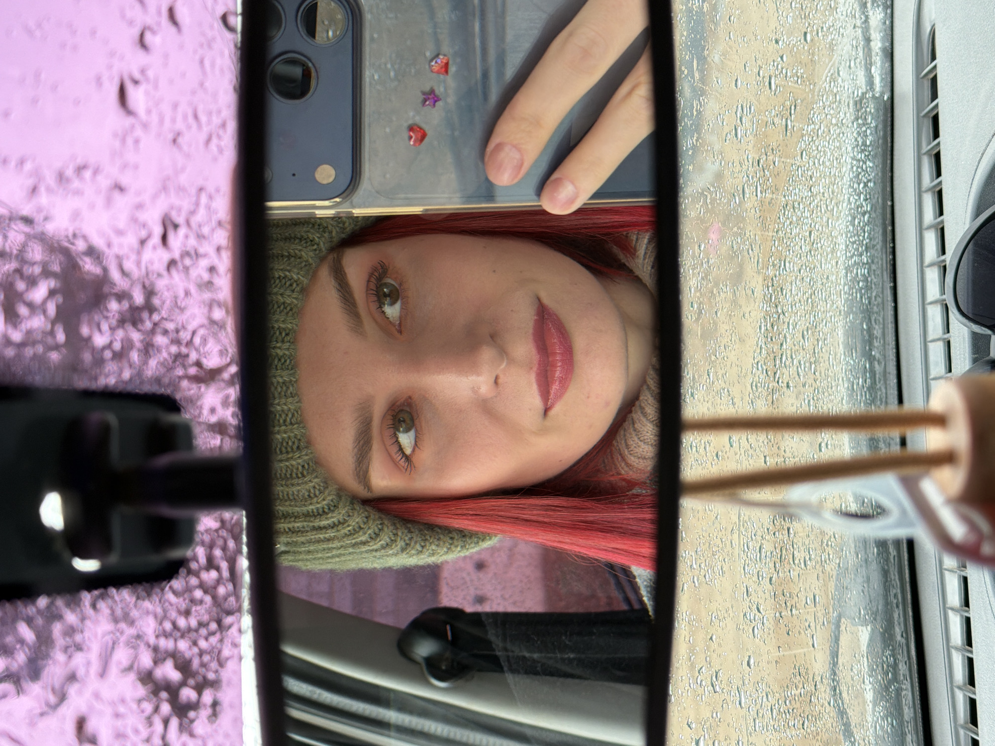 A young woman with prominent red hair and a green knitted beanie hat is captured in a close-up, head-and-shoulders reflection in a car's rearview mirror. She is looking directly towards the camera with a subtle, calm expression and a slight smile, her eyes light green. Her lips are painted with a rosy-red lipstick.
Her right hand, with neatly manicured nails, holds a smartphone (possibly an iPhone given the camera array visible) with a clear case. The phone's case is decorated with small, shiny heart and star-shaped adornments (one red heart, one purple star, another red heart, and possibly a fourth one partially visible).
The scene is set inside a vehicle, likely during daytime, with the view outside the car's windows visible around the mirror's frame. Both the front and side windows appear covered in numerous water droplets, indicating rain or heavy condensation, suggesting damp or wet weather conditions in Chaikovsky, Russia. The background seen through the wet windows on the right side of the image appears light-colored, possibly a dirt road or a distant, muted landscape. On the far right, a car air vent and a part of what looks like an air freshener with two wooden sticks are visible. On the left side, another car interior element, possibly a wiper control, is visible. The overall mood is casual and introspective. No visible text is present in the image.