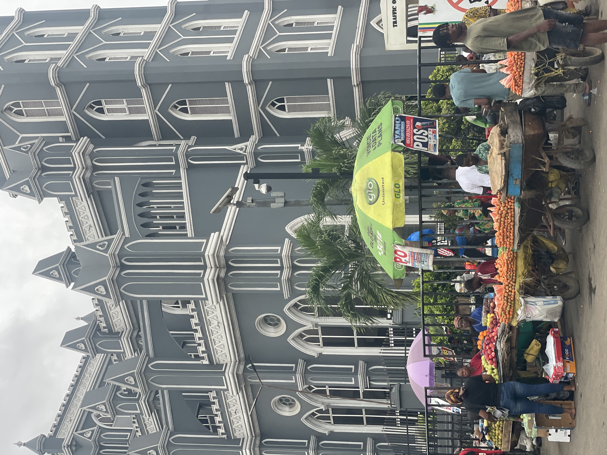 The image shows a street scene in Lagos, Nigeria, with a large grey building with white architectural detailing dominating the left side of the frame. The building has multiple levels and pointed architectural features. A black metal fence runs along the front of the building, separating it from the street.
Along the fence, there are several vendors with carts selling fruits and vegetables, including carrots, apples, and other produce. People are gathered around the carts, presumably shopping. A green and yellow advertising umbrella for "GLO" is visible in the middle of the scene, along with a sign that reads "AYANO RED VENTURES." The vendors seem to be interacting with customers. The street appears busy with activity. A "TRAFFIC OFF" sign is partially visible at the top right.