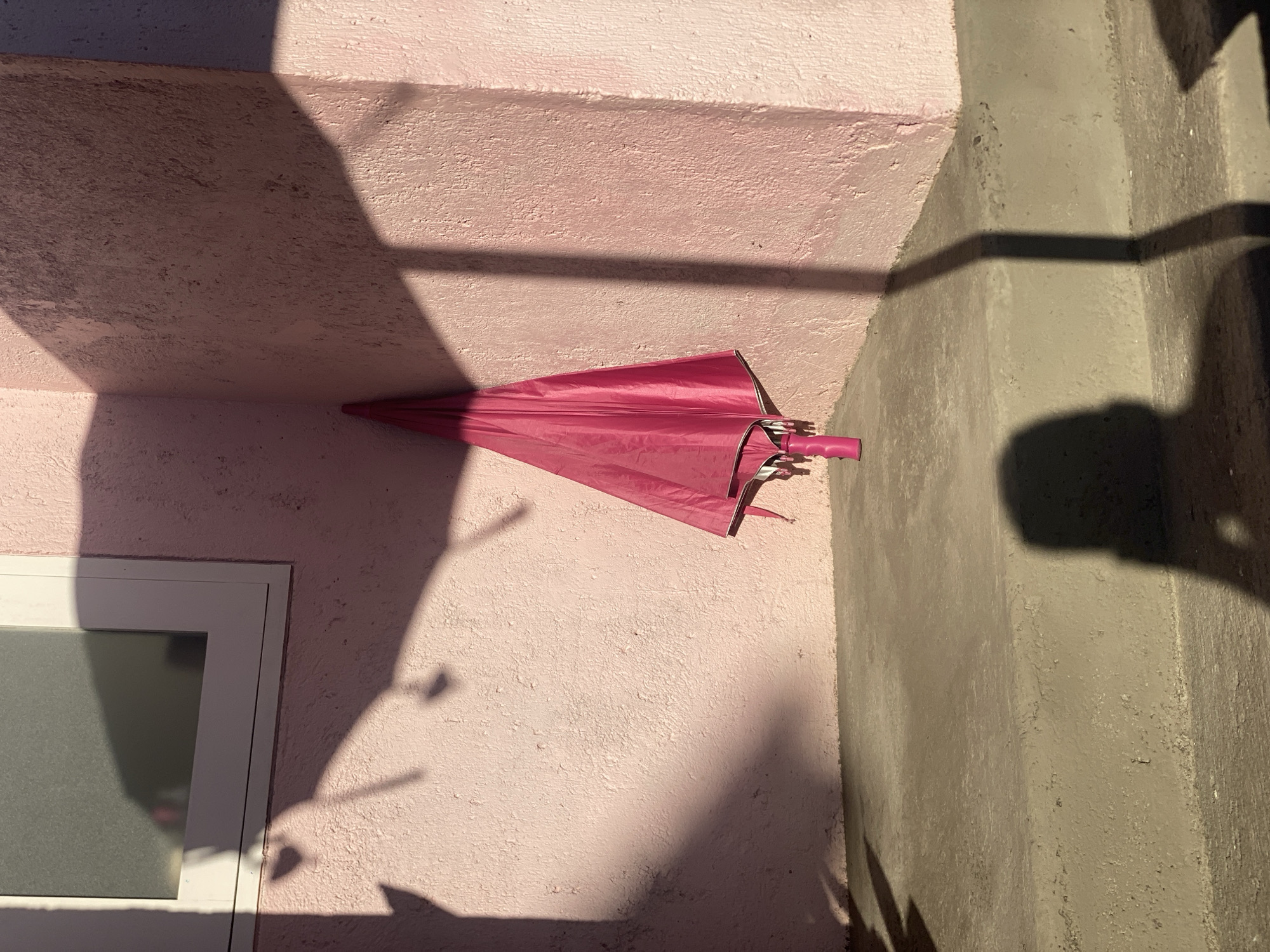 An image shows a pink umbrella leaning against a pink wall in an outdoor setting. The umbrella is closed and its handle is visible at the bottom. Harsh shadows are cast across the wall and a concrete staircase in the foreground. A window with frosted glass is visible on the left side of the image. A shadow of a person is visible on the stairs. The scene appears to be in an urban environment.