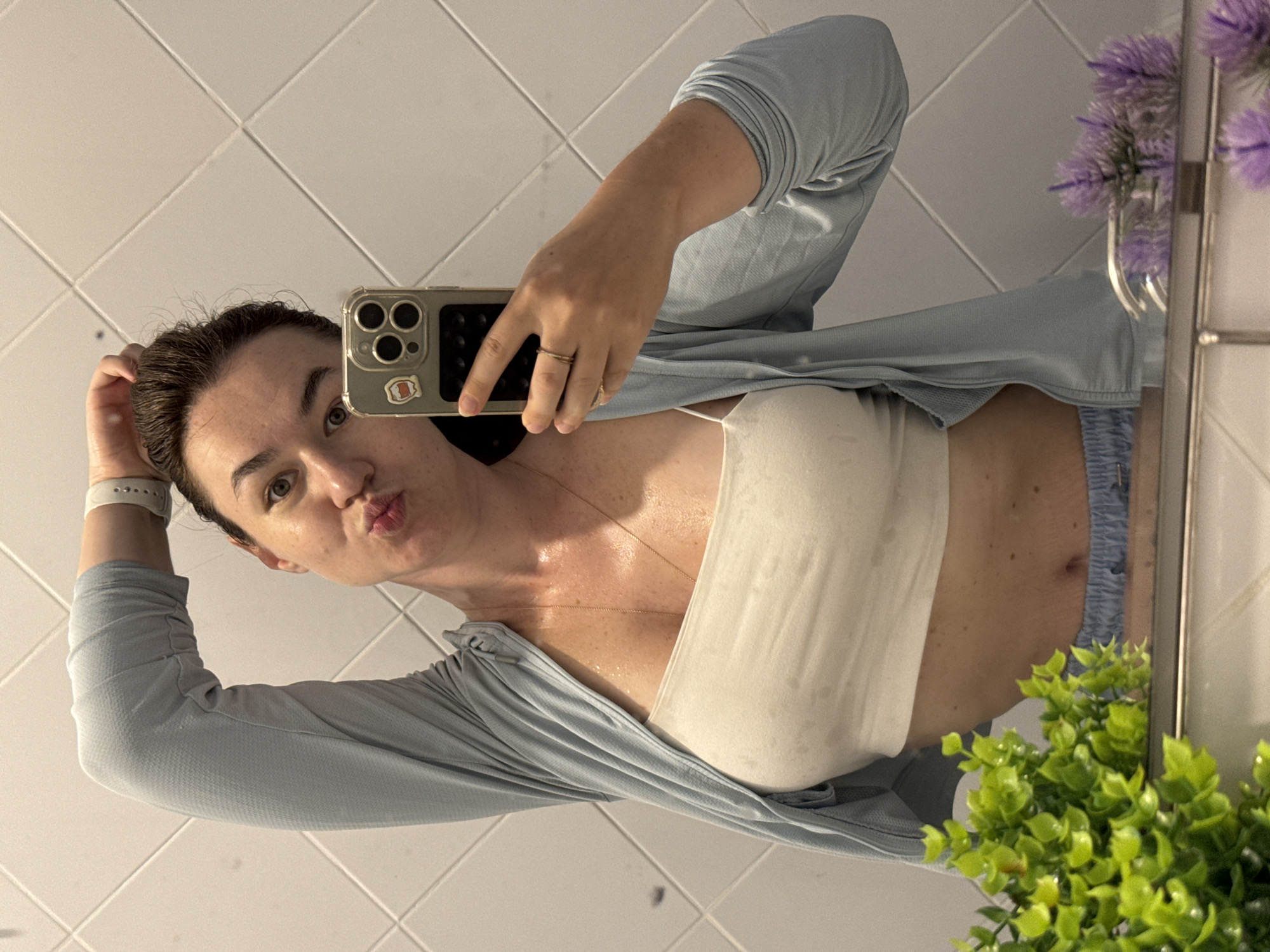 A woman is taking a selfie in a bathroom. She is wearing a light blue cardigan over a white tube top. Her hair is pulled back, and she is making a kissing face. She has a ring on her finger and is holding her phone in her hand. There is a green plant and purple flowers in the foreground. The background is tiled.