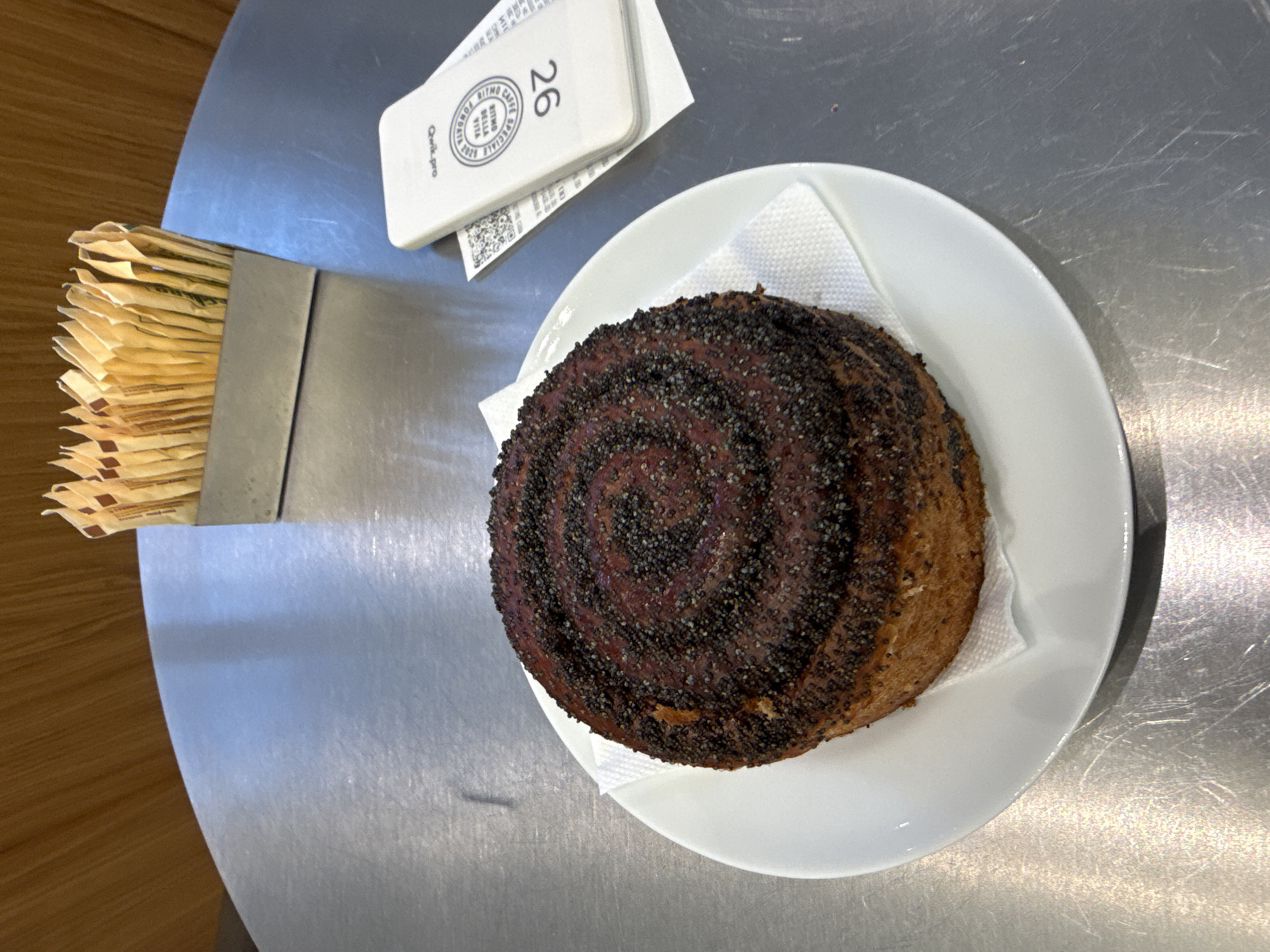 The image shows a top-down view of a table in what appears to be a cafe or restaurant. A round pastry, possibly a sweet roll or bun covered in poppy seeds, sits on a small white plate with a paper napkin. To the left of the plate is a metal container filled with small packets, possibly sugar or sweetener. Above the plate is a white card with the number "26" printed on it, along with some text in Italian "Ritmo Della Vita Fondato Bella Sempre Speciale". The table appears to be metal, and the background includes hints of wood flooring. I can't determine the location of Moscow, Russia based on provided context.