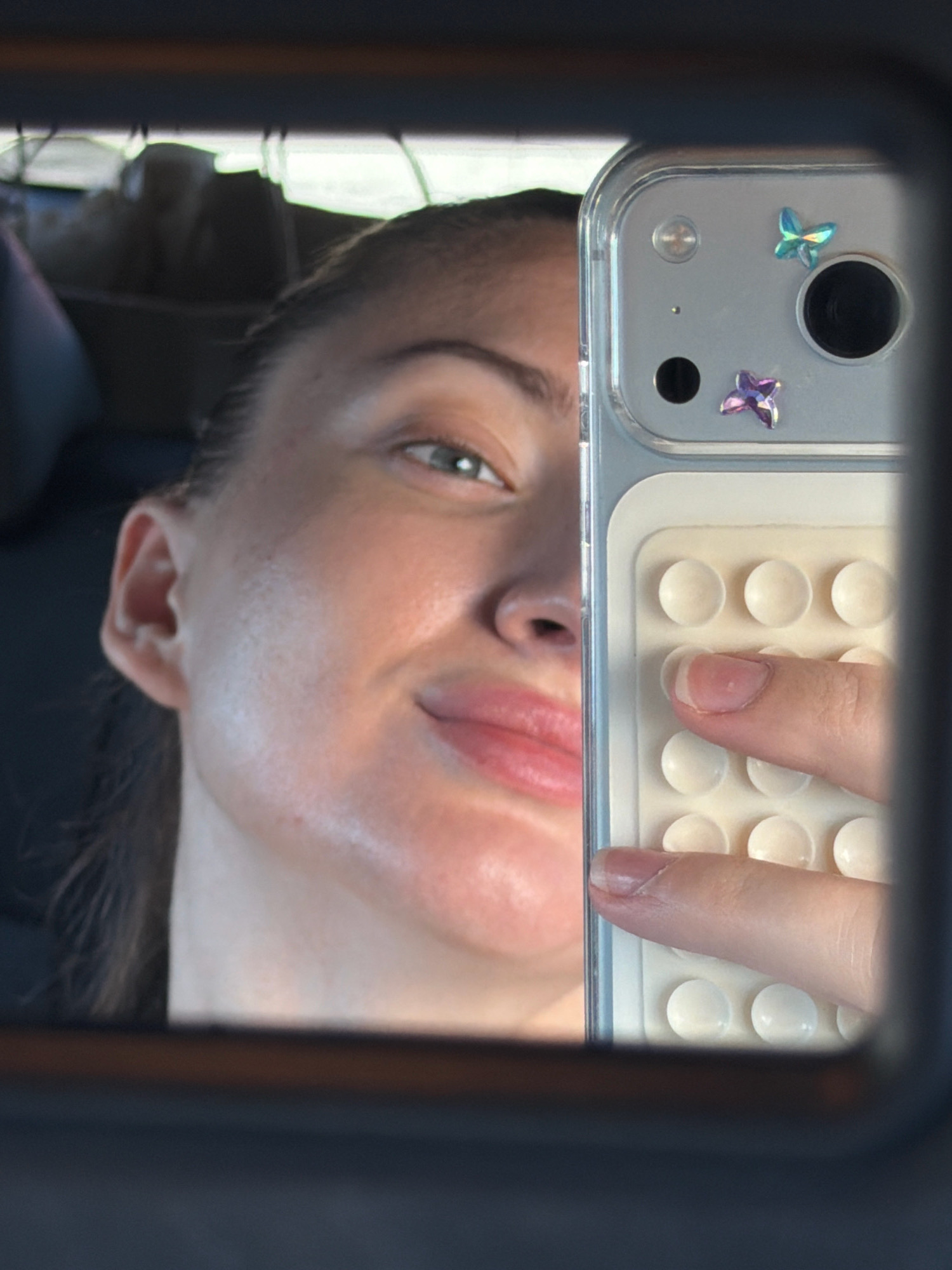 The image is a close-up, distorted selfie taken by a woman reflected in a car mirror. The woman, with dark hair pulled back, is looking towards the camera, with half of her face visible. She has light skin, green eyes, and pink lips. Her right ear is visible. She is holding a smartphone in her right hand, with her fingers obscuring part of the phone's case. The phone case is transparent and has a beige, Lego-like textured back with small white circular protrusions. Two small, iridescent butterfly-shaped stickers, one blue and one purple, are attached to the phone's camera module. The background of the reflection shows the interior of a car, including dark upholstery and some blurry items in the back seat. The lighting suggests it is daytime, possibly in a sunny environment. No specific location within Sharm El Sheikh, Egypt, can be identified from the visual information provided.