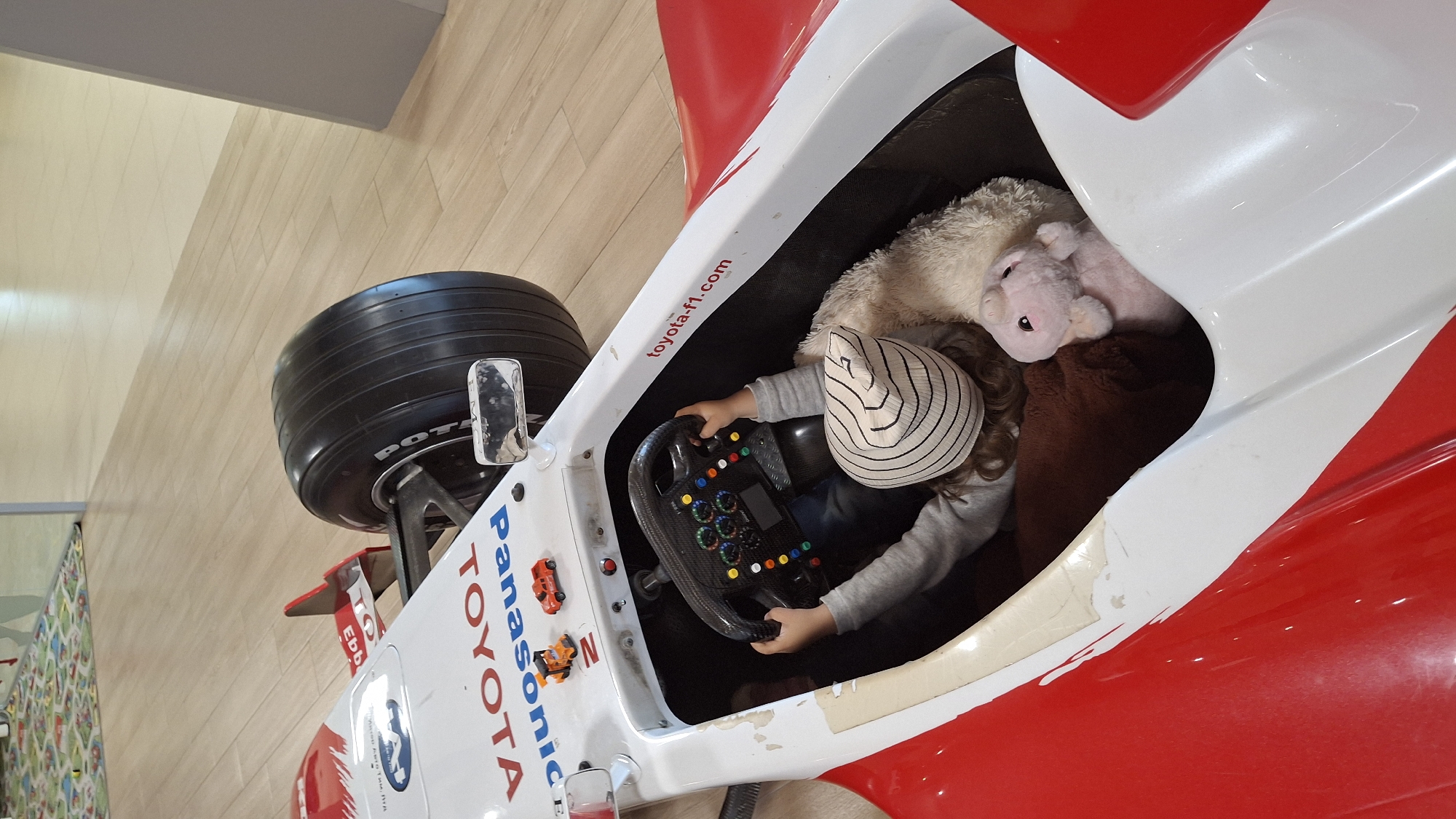 A child is seated in a red and white toy race car, holding onto the steering wheel. The steering wheel is black with colorful buttons and a digital display. The child is wearing a grey long-sleeved shirt and a striped hat. A stuffed pink pig and a fluffy white toy are also in the car with the child. The car has "TOYOTA" and "Panasonic" logos on its exterior, and "toyota-f1.com" is written on the side. The car is on a wooden floor.