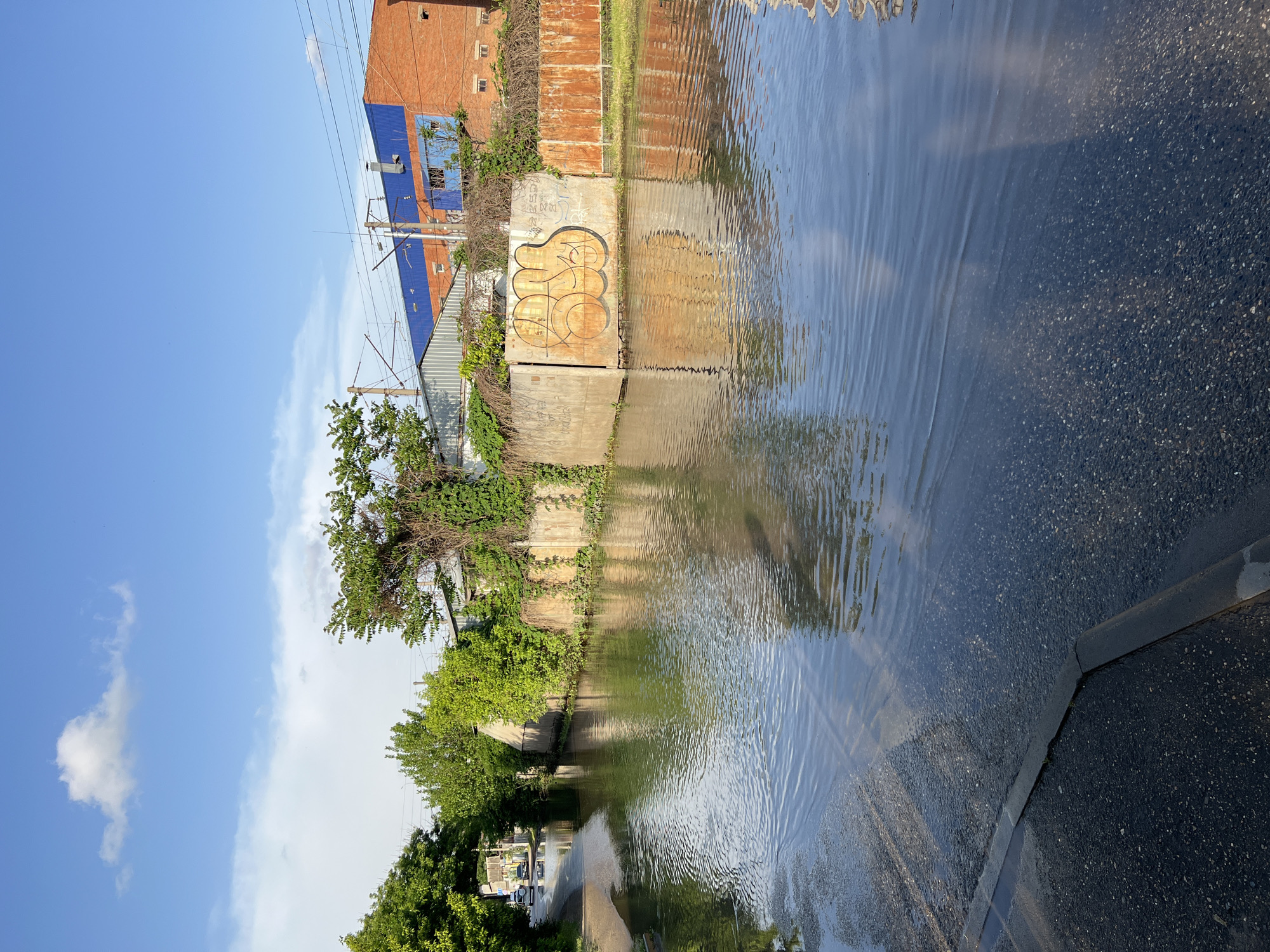 The image shows a flooded street in Krasnodar, Russia. The water level reaches the sidewalk, partially submerging the pavement. Buildings and structures are visible along the edge of the water, including a brick building with blue roofing and a concrete wall with graffiti. Vegetation grows along the water's edge. The sky is visible with some clouds. The water's surface is rippled, reflecting the surrounding environment.