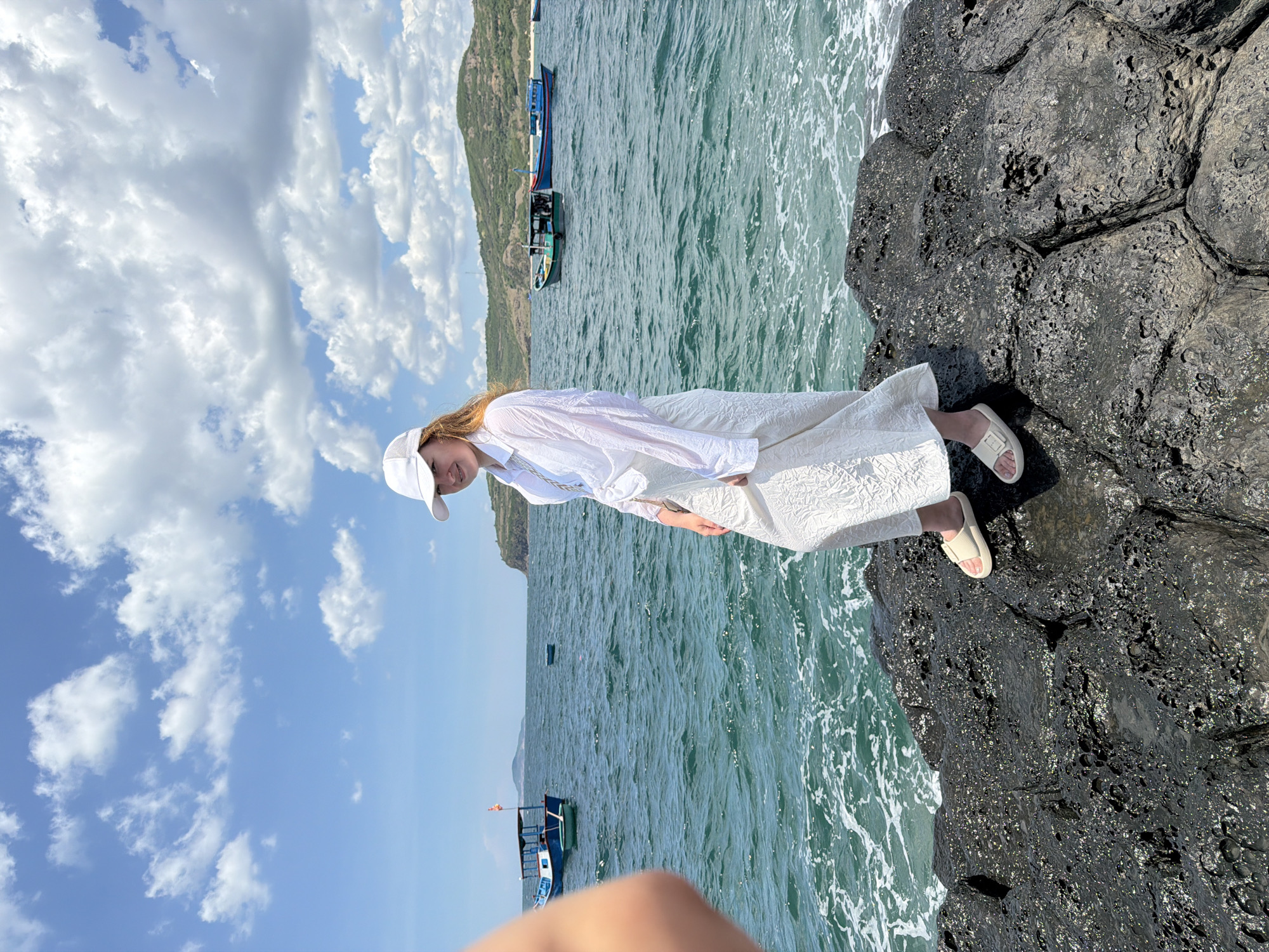 A young woman, facing the viewer and smiling, stands on a rocky outcrop by the sea. She is wearing a white cap, a white open-necked shirt, and a textured white maxi skirt, with light-colored slides on her feet. Her left arm is visible in the foreground, blurred and out of focus. The sea is a vibrant turquoise, with small waves lapping at the rocks. Several fishing boats are visible on the water, and a hilly coastline can be seen in the distance. The sky is blue with scattered white clouds. The location appears to be a coastal area in Phú Lương, Vietnam.
