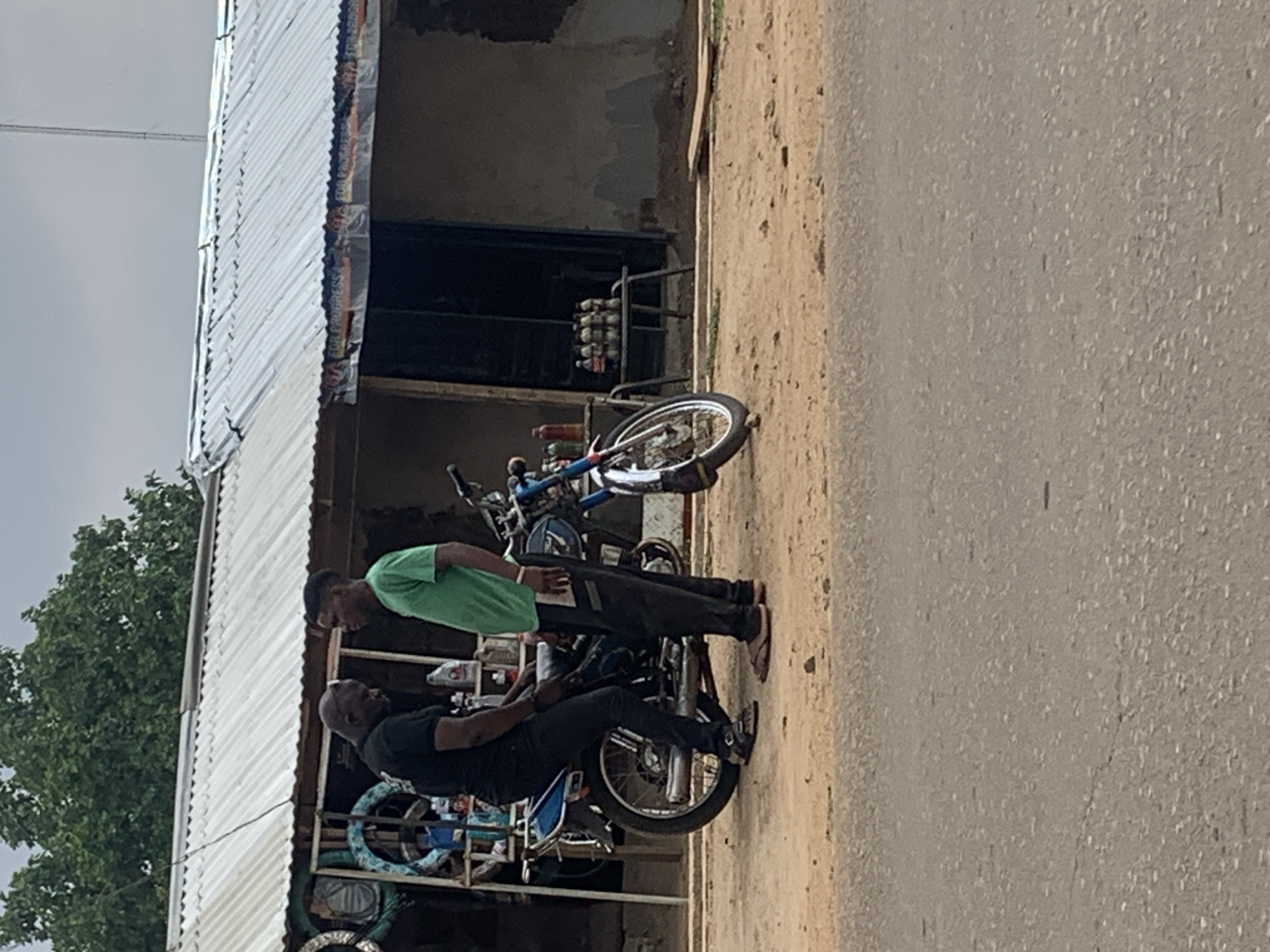 The image, taken in Maigizo, Nigeria, depicts an outdoor street scene with two men, motorcycles, and a building under an overcast sky.
The primary subjects are two men and several motorcycles. One man, wearing a green t-shirt and dark trousers, stands upright, looking down towards the other man. The second man, dressed in a black t-shirt and dark trousers, is seated on a blue and silver motorcycle, facing the first man. They appear to be engaged in a conversation or an exchange. Another motorcycle is partially visible behind them, and a bicycle leans against the structure.
Behind the men and vehicles is a single-story building featuring a prominent corrugated metal roof. The walls below the roof are a light, muted color, possibly concrete or stucco. A metal rack holds several small, clear plastic bottles, likely containing liquids such as water or oil, near the entrance of the building. Another small bottle is visible on a surface closer to the men.
The setting includes a dirt path directly in front of the building, which transitions into an asphalt road on the right side of the frame. Lush green foliage from a tree is visible on the far left, extending towards the sky. The sky itself is cloudy and overcast, indicating daytime but without direct sunlight.
A notable detail is visible text along the edge of the corrugated roof, which appears to be part of a banner or advertisement. The text clearly reads "FOR PROGRESS" repeated, suggesting a political slogan or a business motto. The overall atmosphere is mundane and typical of a local street or market area.