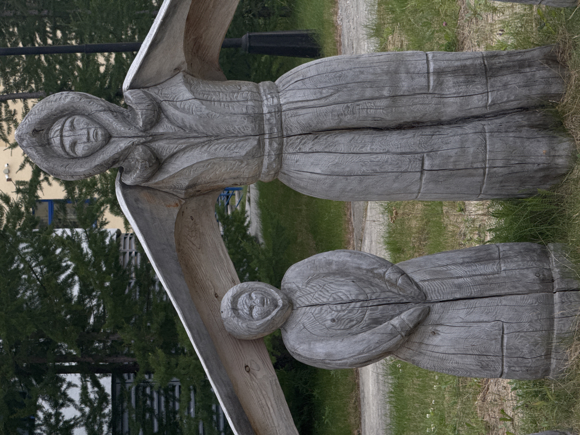 The image displays two weathered wooden sculptures of figures dressed in traditional clothing, possibly resembling indigenous attire. The sculptures are standing upright in a grassy outdoor area, with a gravel path visible behind them. A modern building and evergreen trees form the background. The sculpture on the right is taller and more prominent, with its arms outstretched, supporting a large, angled wooden beam. The sculpture on the left is smaller and positioned in front of the larger one, appearing to be a child or a companion figure. The wood shows significant signs of age and weathering, with visible grain and cracks. The context provided by the user suggests this scene is located in Salekhard, Russia. The overall impression is one of outdoor art or a cultural display.