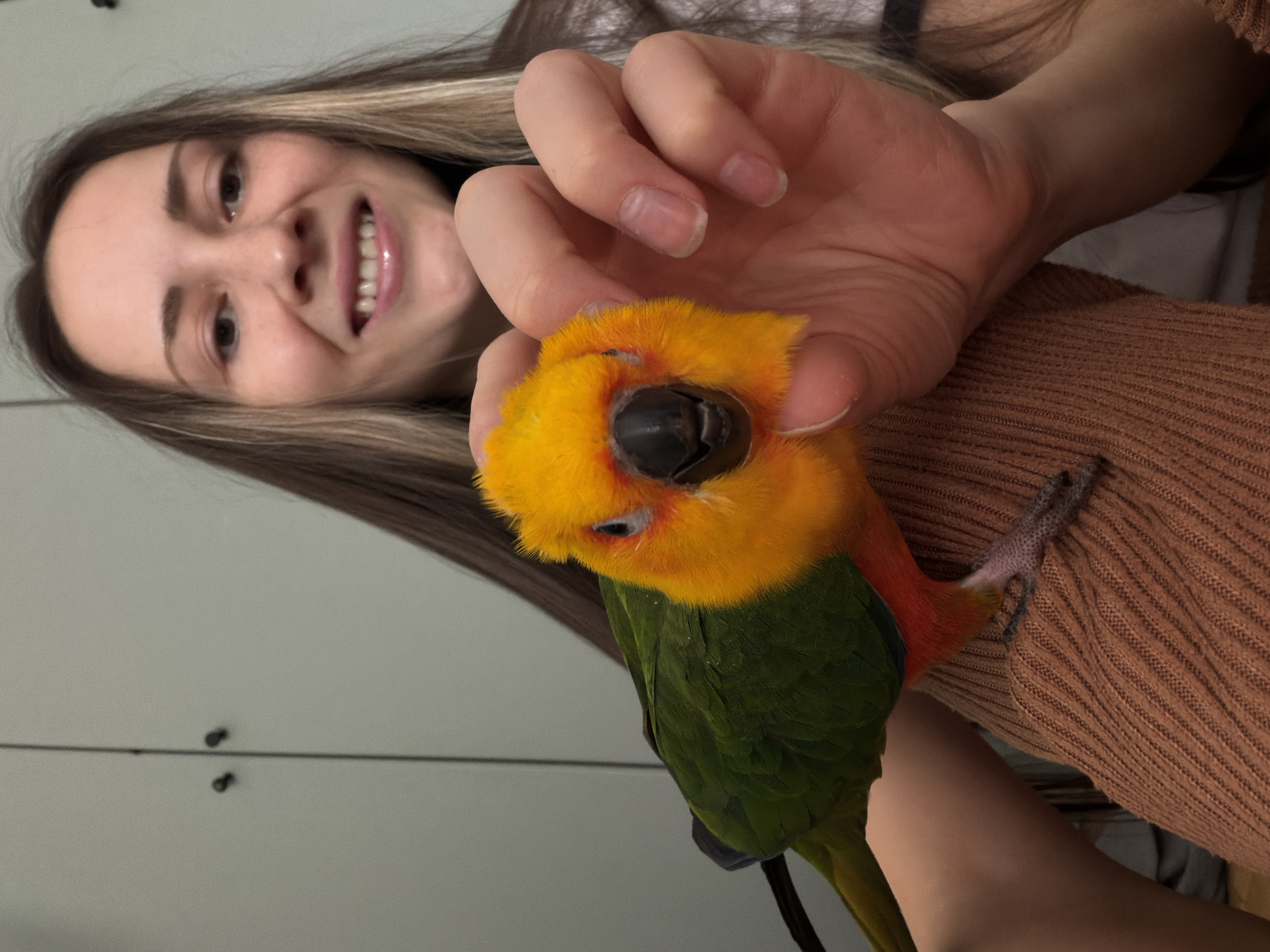 A woman with shoulder-length light brown hair and a wide smile is holding a colorful parrot. The parrot has a yellow head, green body, and orange markings around its neck. The woman is wearing a brown sweater. The background is a light gray wall with a door. The location in Khimki, Russia, cannot be determined from the image.