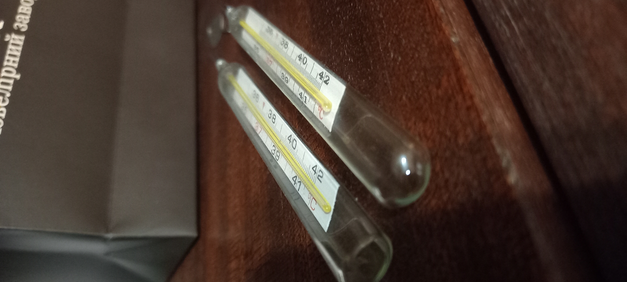 This image captures a still life featuring two glass thermometers resting on a dark, textured surface, likely leather or wood. To the left, a dark box or bag is partially visible.
The thermometers are clinical mercury or gallium thermometers, characterized by their slender glass tubes and visible metallic columns. Both display a temperature scale ranging from approximately 36°C to 42°C, with prominent markings at 37, 38, 39, 40, 41, and 42 degrees Celsius. A red indicator line is visible at 37°C on both, typically signifying normal human body temperature. The measuring liquid inside appears to be below 37°C in both, suggesting they are at room temperature or have been reset.
On the dark box or bag to the left, partially visible white text in Cyrillic reads "ювелірний заво", which translates to "jewelry factory" (part of the word "завод" - factory - is cut off). This suggests the box might be packaging from a jewelry manufacturer.
There are no people or specific activities depicted in the image. The setting appears to be indoors, under artificial lighting, given the reflections on the thermometers and the overall subdued lighting. The image provides no direct cues regarding the time of day or weather. The provided context indicates the location is Kryvyi Rih, Ukraine.