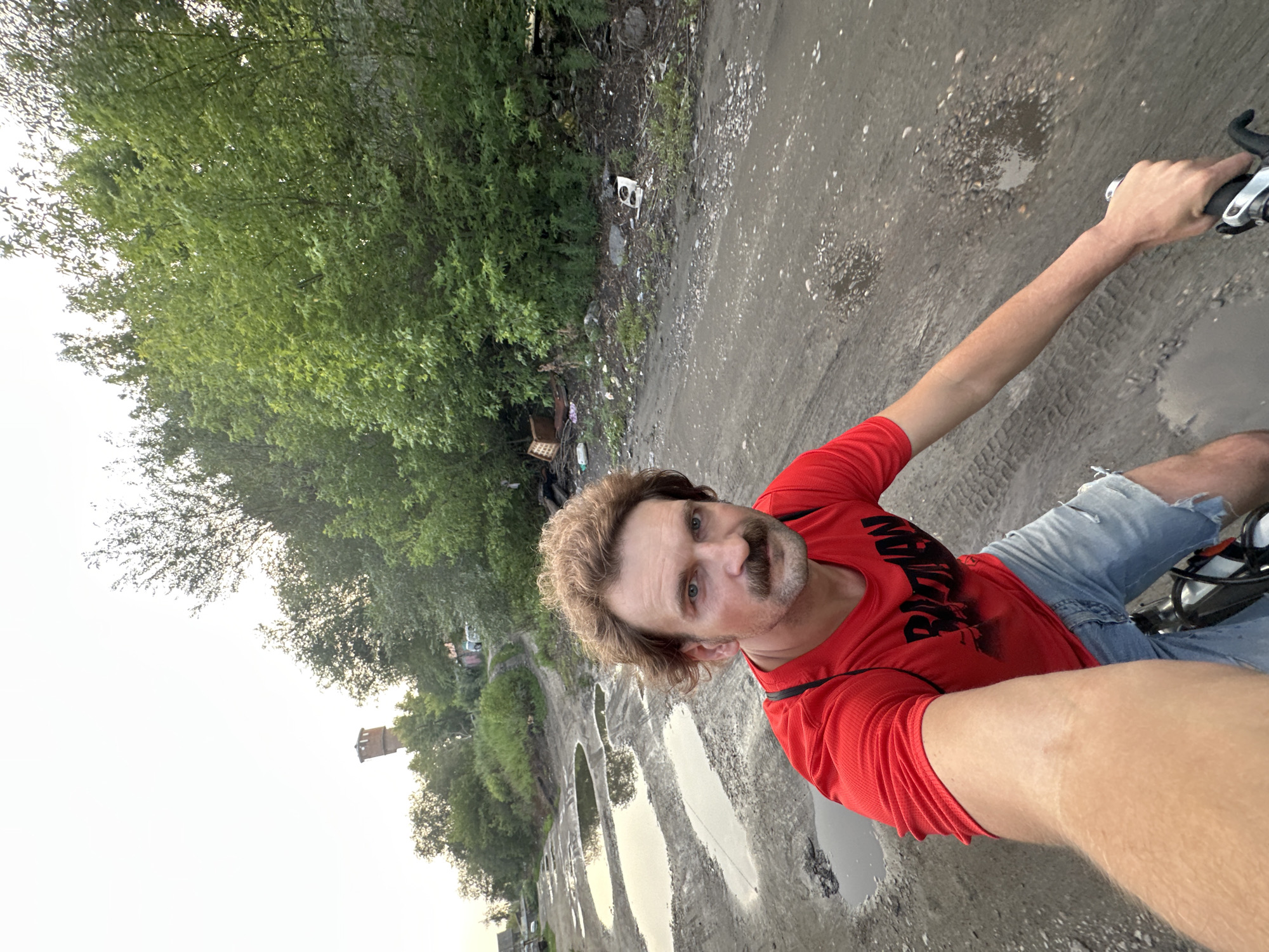 A man with a mustache and wavy brown hair, wearing a red t-shirt with a black graphic and denim shorts, rides a bicycle on a dirt road with puddles. The road is lined with trees and greenery on the right, and on the left there is more greenery and a water tower in the distance. There is debris and trash scattered along the side of the road. The sky is overcast. The scene appears to be in an industrial or neglected area of Biysk, Russia.