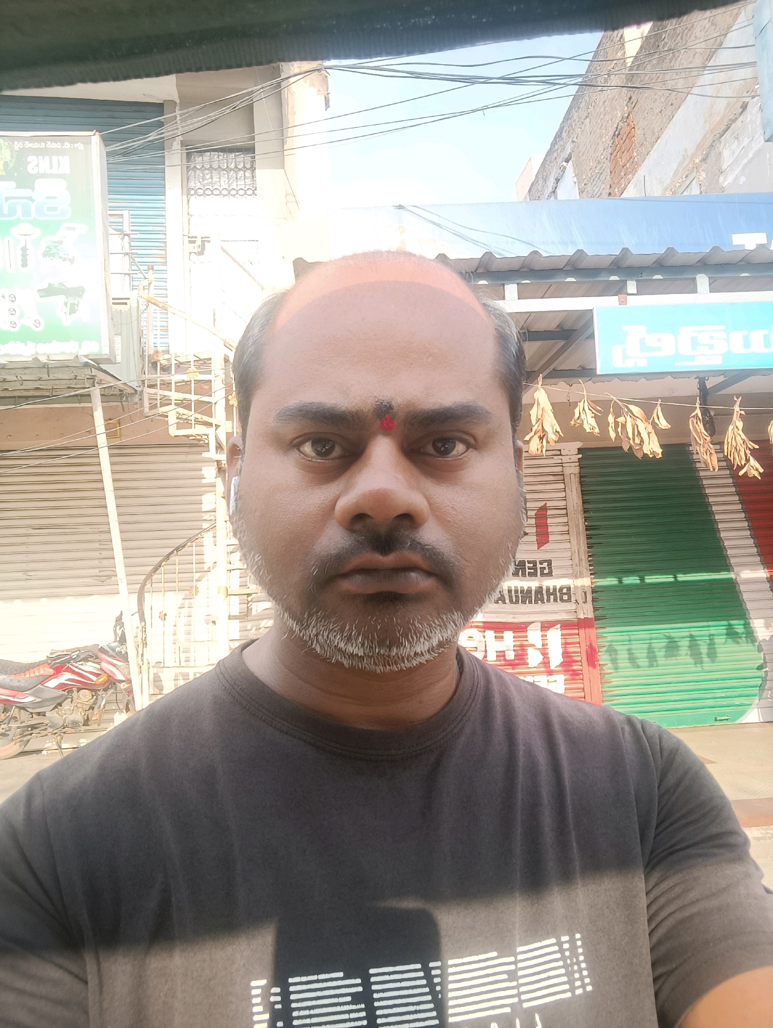 A man is captured in a close-up selfie in Kadiri, India. He is looking directly at the camera with a neutral expression. He has a dark complexion, a receding hairline, and a salt-and-pepper beard and mustache. A red "bindi" is visible on his forehead. He is wearing a dark t-shirt. In the background, there are buildings with closed shutters, some signs with text, and a motorcycle parked to the left. The overall scene suggests an outdoor setting in a commercial or residential area of the city.