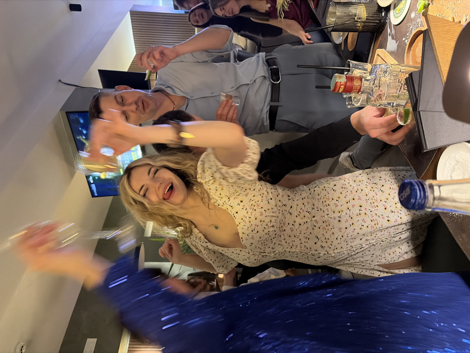 The image depicts an indoor celebratory gathering within a modern bar or karaoke lounge in Budva, Montenegro. In the central foreground, a blonde woman with wavy hair, wearing a white floral-patterned dress with puffed sleeves, smiles broadly with her tongue extended, actively raising a glass in her right hand, suggesting dancing or a toast. To her left, a blurred figure in a shimmering blue sequined garment also raises a glass. To the right of the central woman, a man in a light blue collared shirt and grey trousers holds a shot glass in his left hand and a lime wedge in his right, appearing to prepare for a drink. The immediate foreground shows a dark table surface laden with various items: a clear bottle with a blue cap, a tequila bottle, multiple shot glasses containing liquid and lime wedges, dark tumblers with straws, coasters, and scattered salt or sugar. An arm in a black long-sleeved top is visible on the table, interacting with the shot glasses. In the background, additional individuals are present, some smiling, contributing to a lively atmosphere. Two wall-mounted flat-screen televisions display Cyrillic text, including "КАРАОКЕ КАРТА" (Karaoke Menu). The interior features contemporary lighting and wall paneling, with vertical wooden slats visible in the background.